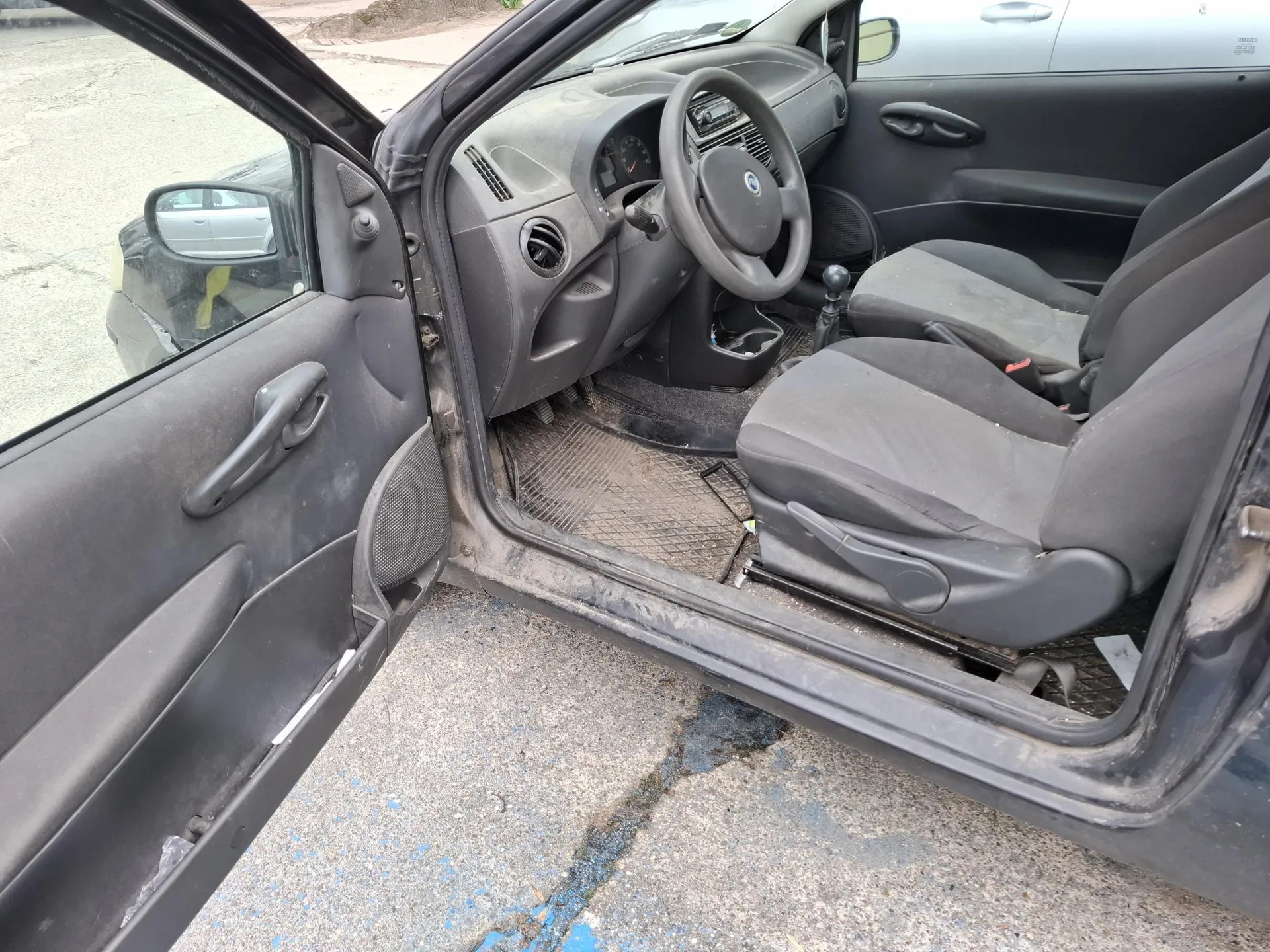 Wnętrze samochodu z otwartą lewą przodem drzwiami. Widoczne są czarne siedzenia, kierownica z logo, deska rozdzielcza z licznikami, dźwignia zmiany biegów oraz brudne dywaniki podłogowe. Poza samochodem widoczna jest asfaltowa powierzchnia oraz fragment innego pojazdu w lusterku bocznym.