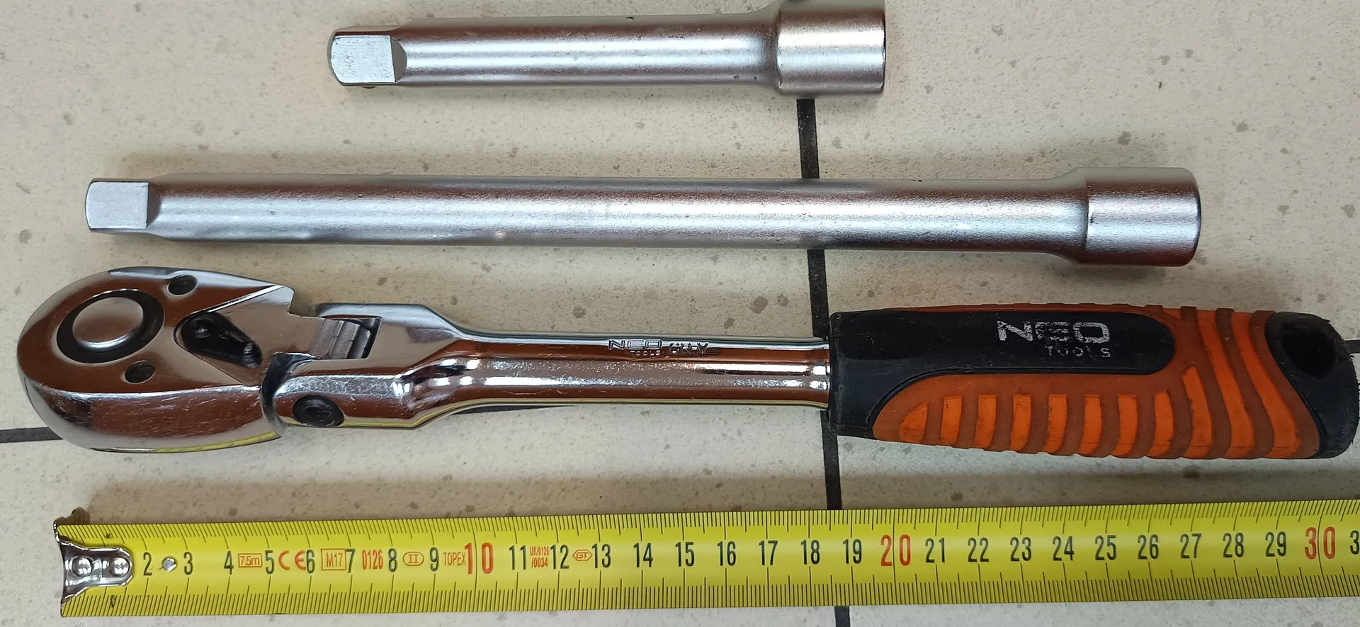 Na obrazie widoczne są narzędzia ułożone na jasnej powierzchni. Po lewej stronie znajduje się żółta taśma miernicza z czarnymi i czerwonymi oznaczeniami od 2 do 30. Obok niej leży klucz nasadowy z pomarańczowo-czarną rączką, na której widnieje napis 'NEO TOOLS'. Obok klucza znajdują się dwie metalowe nasadki: jedna dłuższa, druga krótsza, obie z sześciokątnymi końcówkami. Tło stanowi jasna powierzchnia z ciemnymi liniami.