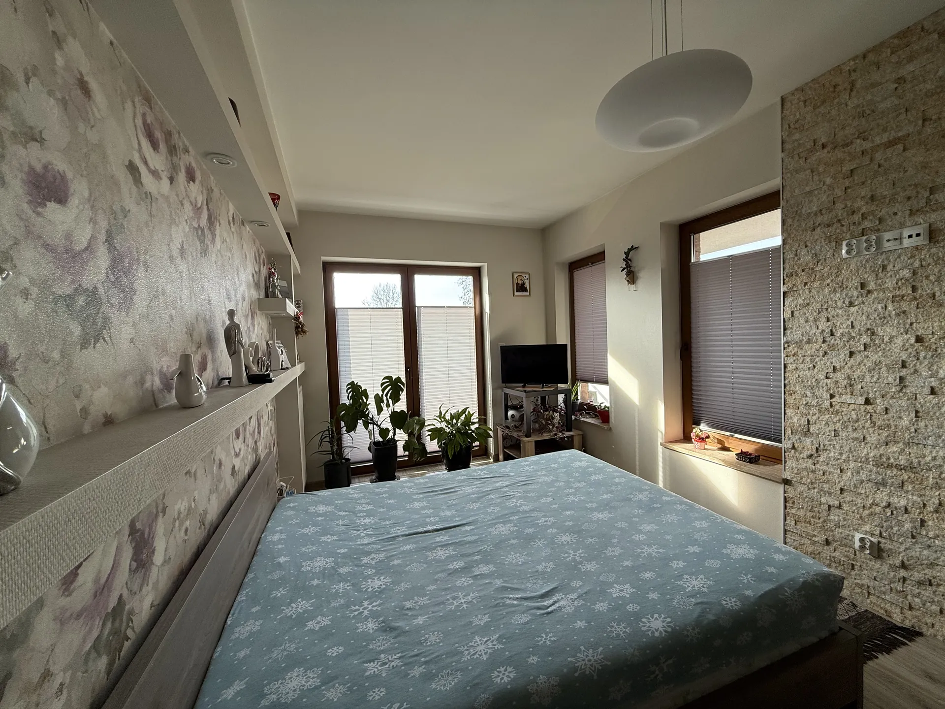 Sypialnia z łóżkiem przykrytym niebieską pościelą z białymi wzorami płatków śniegu. Po lewej stronie ściana z kwiecistą tapetą i półką z dekoracyjnymi wazonami oraz figurkami. Po prawej ściana z teksturą kamienia i gniazdami elektrycznymi. Trzy okna z roletami: duże drzwi przesuwne z oknami w tle oraz dwa mniejsze po prawej stronie. Przy drzwiach przesuwnych stoją doniczki z roślinami. W kącie telewizor na stojaku. Sufit z okrągłym wiszącym lampą. Podłoga z drewnianych desek i dywanik przy łóżku.