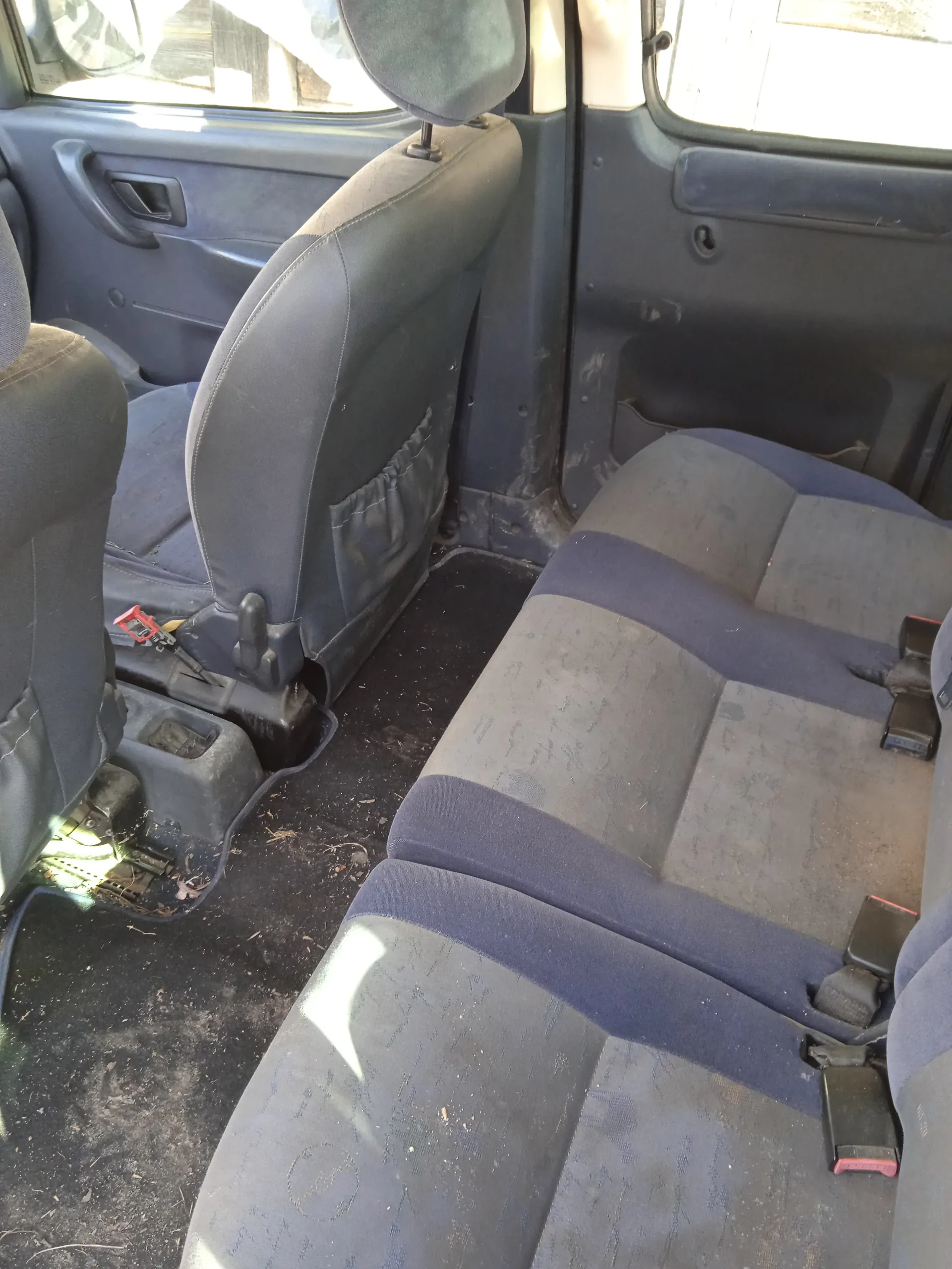 Wnętrze samochodu z widokiem na tylne siedzenia. Siedzenia są w kolorze ciemnoniebieskim i szarym, z widocznymi śladami brudu i zużycia. Widoczne są pasy bezpieczeństwa z czerwonymi przyciskami, podłoga pokryta brudem i drobnymi szczątkami. Drzwi samochodu są otwarte, pokazując fragmenty paneli drzwiowych. W tle widoczne są przednie siedzenia z zagłówkami oraz fragmenty okien.
