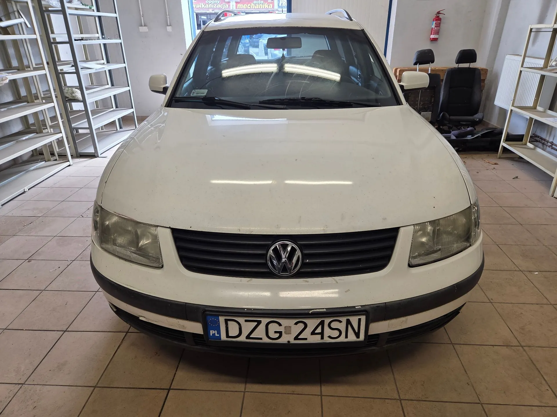 Biały samochód marki Volkswagen Passat w nadwoziu kombi, zaparkowany wewnątrz warsztatu. Widoczny jest przód pojazdu z tablicą rejestracyjną o numerze DZG 24SN. W tle znajdują się białe regały magazynowe, gaśnica oraz fotele samochodowe.