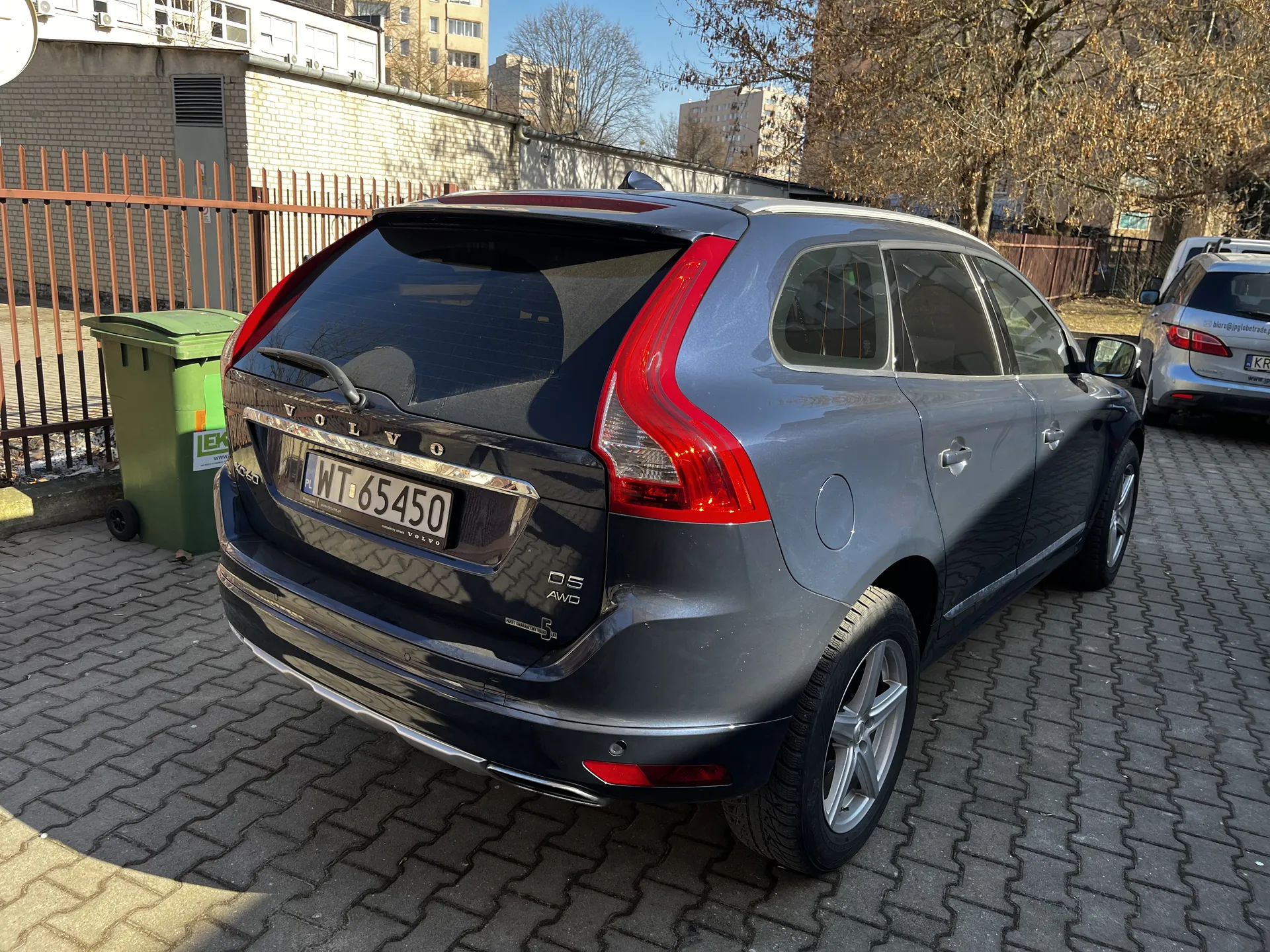 Srebrny samochód Volvo typu kombi zaparkowany na brukowanej powierzchni. Widoczny tył pojazdu z tablicą rejestracyjną WT 65450, czerwonymi światłami tylnymi i napisem VOLVO. Po lewej stronie zielony pojemnik na śmieci z naklejką LEK oraz pomarańczowe ogrodzenie. W tle budynki mieszkalne, drzewa bez liści i jasne niebo. W oddali drugi samochód z częściowo widoczną tablicą rejestracyjną KR.