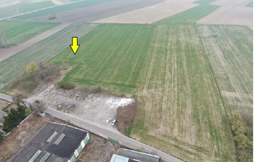 Zdjęcie lotnicze przedstawiające obszar rolniczy z polami o różnych odcieniach zieleni i brązu. W dolnej części obrazu widoczny jest betonowy plac z zaparkowanymi dwoma białymi samochodami oraz fragmenty budynków przemysłowych z ciemnymi dachami. Na zielonym polu w górnej lewej części zdjęcia znajduje się żółta strzałka skierowana w dół.