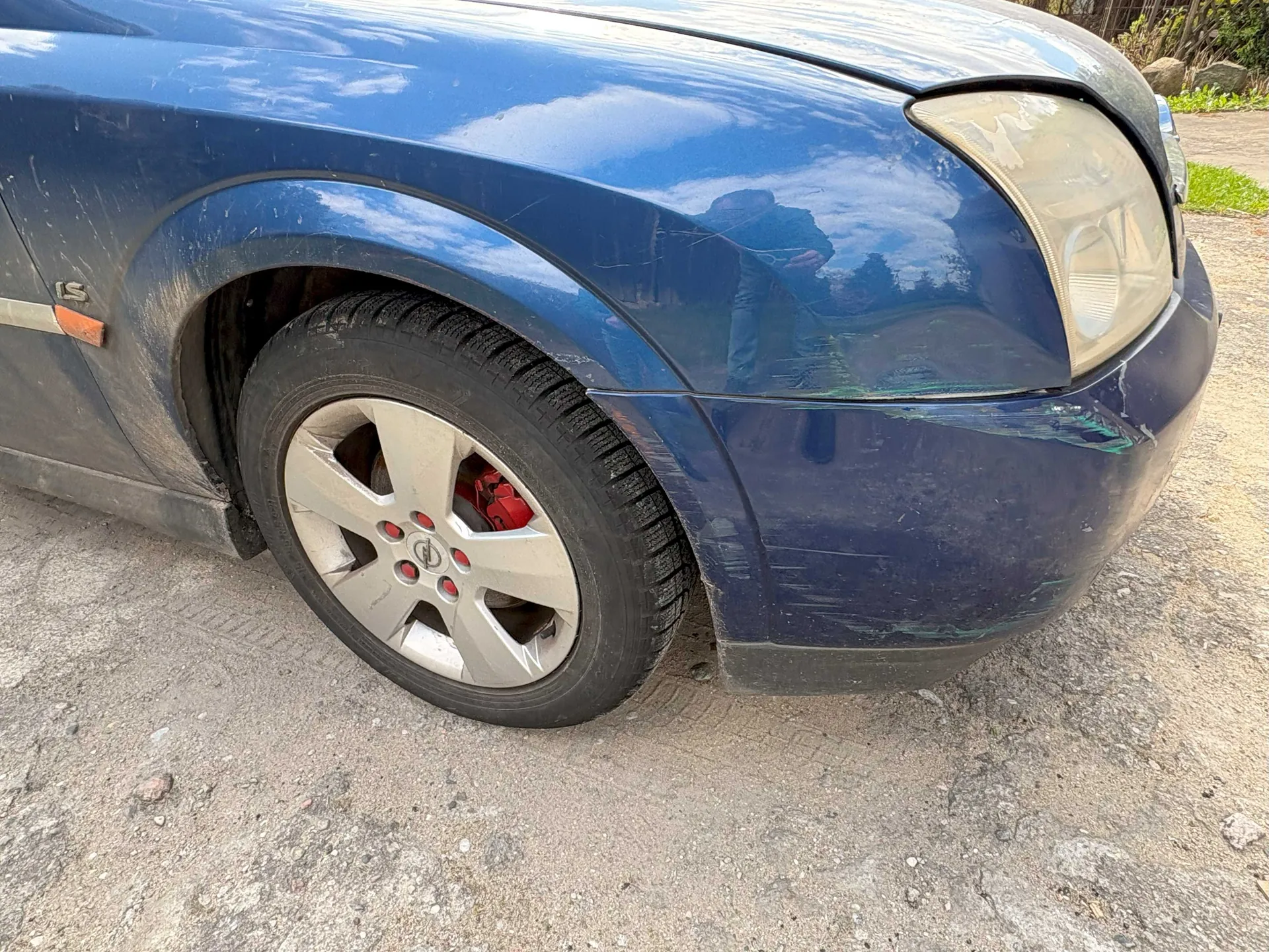 Przednia prawa strona niebieskiego samochodu z widocznym reflektorem, kołem z srebrnym felgą i czerwonymi zaciskami hamulcowymi. Na boku nadwozia widoczny napis 'LS' oraz pomarańczowy odblask. Samochód stoi na drodze z żwiru, w tle trawa i kamienie. Nadwozie pokryte jest kurzem, a na karoserii odbijają się niebo i drzewa.