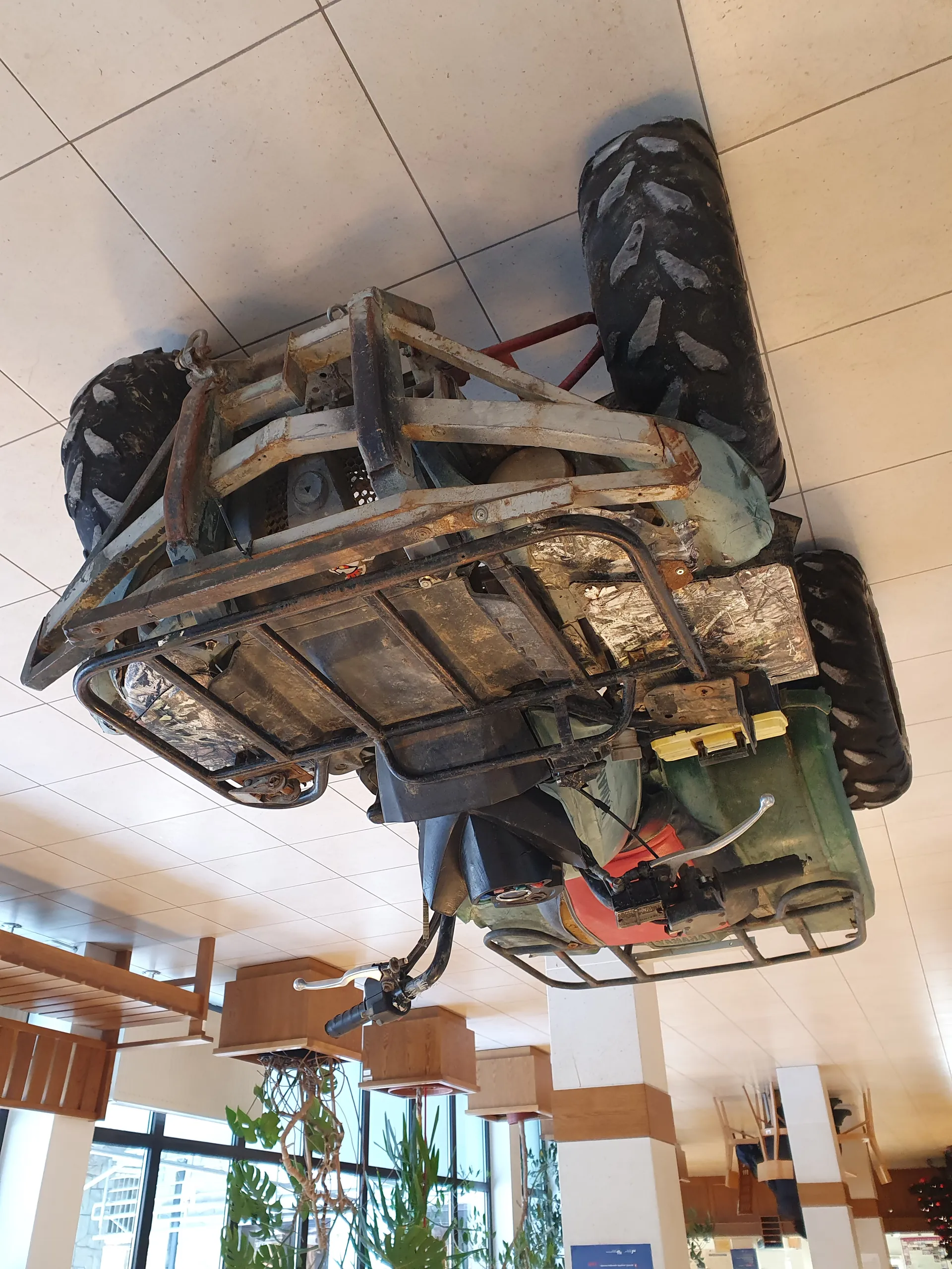 Zardzewiały quad z dużymi oponami umieszczony w pomieszczeniu o podłodze z białymi płytkami. W tle widoczne są drewniane krzesła, donice z roślinami oraz duże okna. Quad posiada metalową ramę, widoczne ślady zużycia i rdzy, a także żółte elementy. Pomieszczenie przypomina hol lub przestrzeń wystawienniczą.