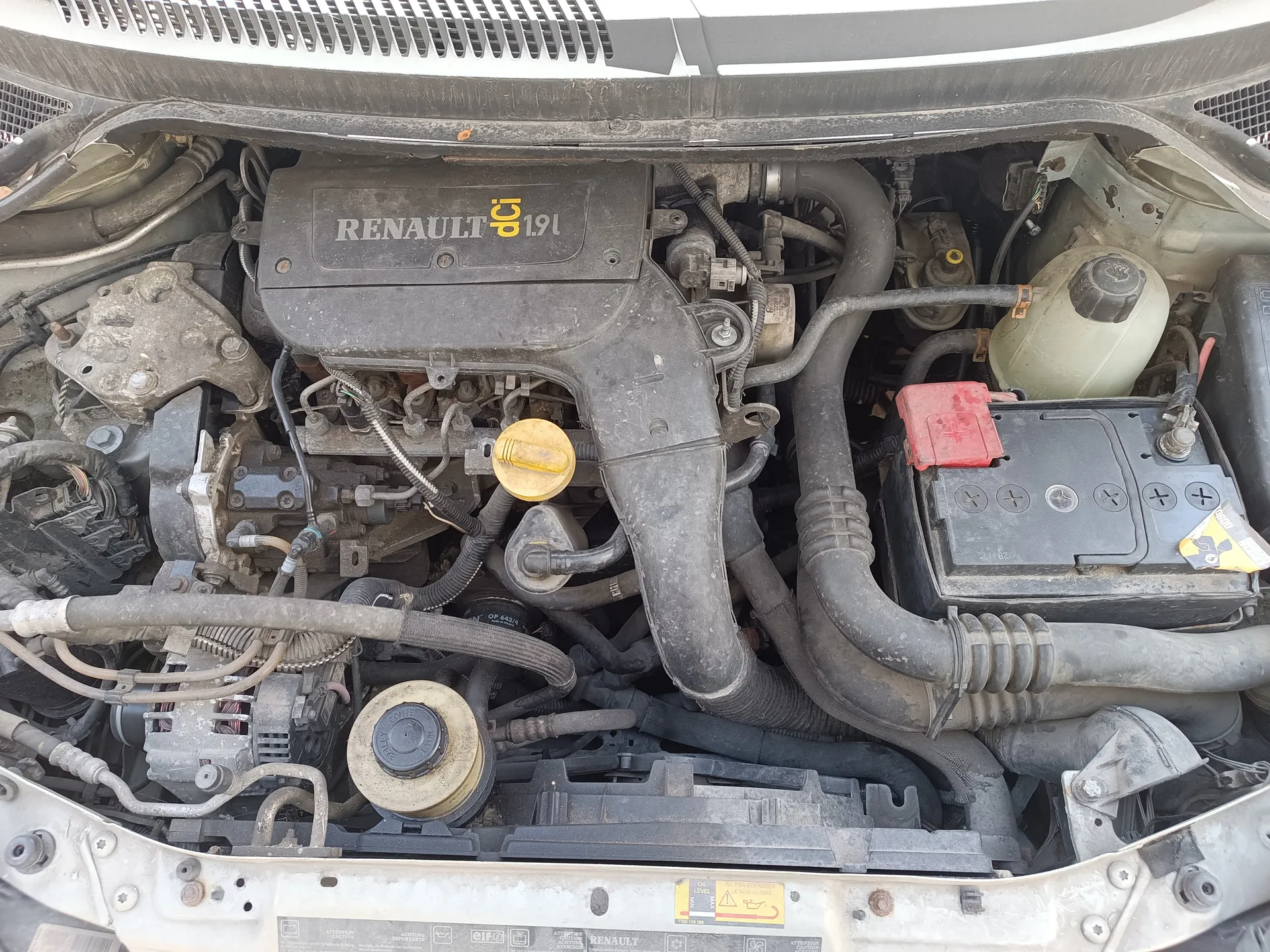 Silnik samochodu marki Renault z widoczną pokrywą z napisem RENAULT dCi 1.9l. W centralnej części znajduje się żółty korek do oleju, po prawej stronie czarna bateria z czerwonym kablem oraz zbiornik chłodniczy. Wokół widoczne są różne przewody, węże i elementy mechaniczne. Na dole obrazu znajduje się etykieta z napisami i symbolami, w tym logo Renault oraz ostrzeżeniami.