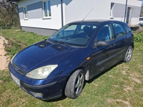 Ciemnoniebieski samochód osobowy typu hatchback marki Ford Focus zaparkowany na trawie. Pojazd znajduje się pod kątem, z przodem skierowanym w lewo. W tle budynek mieszkalny z białymi i szarymi ścianami, oknami oraz drzwiami. Trawa na podwórku jest zielona z widocznymi plamami ziemi. Na przednim zderzaku widoczna tablica rejestracyjna.