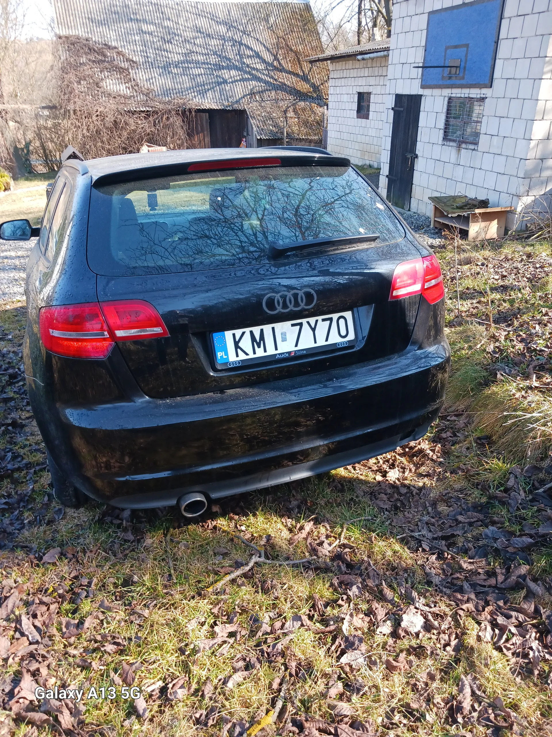 Czarny samochód marki Audi z tablicą rejestracyjną KMI 7Y70 zaparkowany na trawie z suchymi liśćmi. W tle budynek z białymi płytkami i niebieską tablicą koszykówki. W prawym dolnym rogu widoczny napis 'Galaxy A13 5G'.