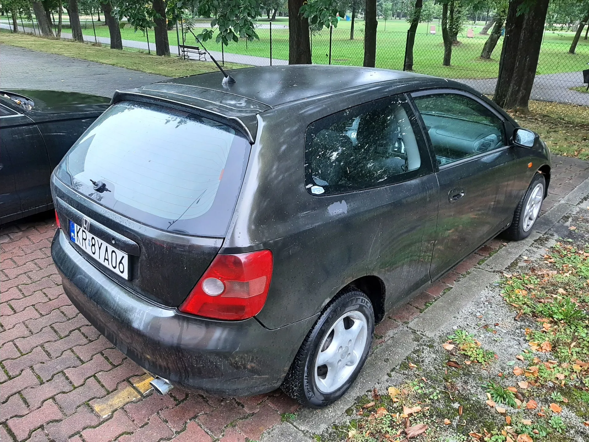 Czarny, brudny samochód osobowy typu hatchback zaparkowany na kostce brukowej obok trawnika i drzew. Widoczna jest tylna część pojazdu z tablicą rejestracyjną o numerze KR 8YA06. Samochód jest pokryty warstwą kurzu i błota.