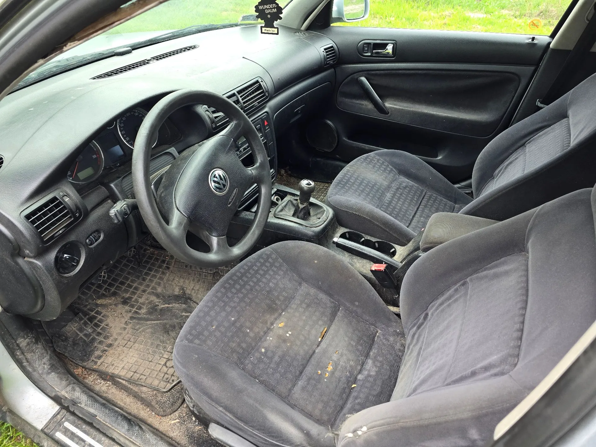 Wnętrze samochodu marki Volkswagen z widokiem na przednie siedzenia, deskę rozdzielczą i kierownicę. Tapicerka siedzeń jest ciemnoszara w drobną kratkę i widoczne są na niej zabrudzenia. Kierownica posiada logo Volkswagena w centralnej części. Wnętrze jest zakurzone, a na podłodze znajduje się gumowa mata.