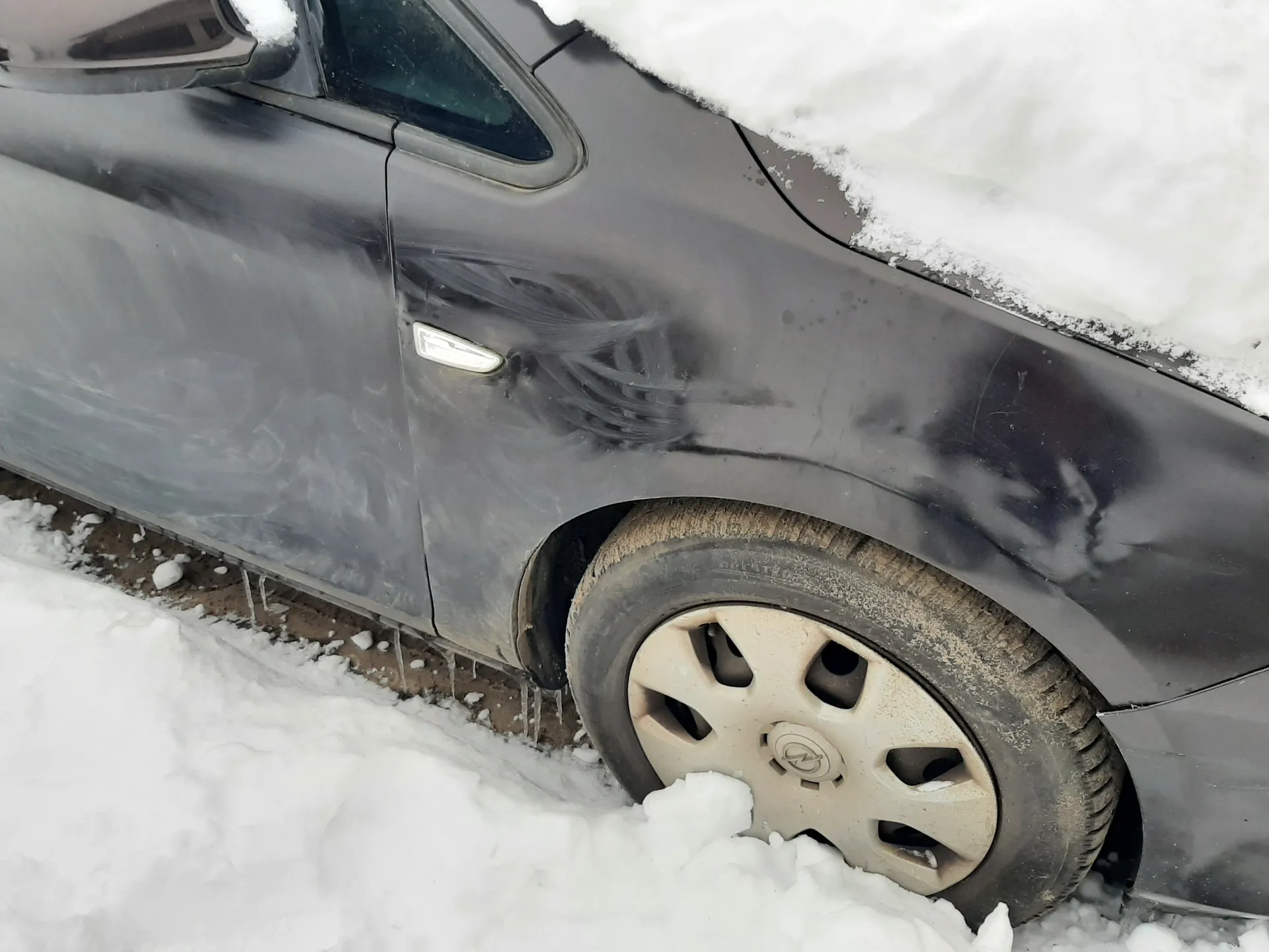 Ciemnoszary samochód pokryty śniegiem na dachu i kołach. Widoczne zadrapania na drzwiach, brudna opona z białą felgą. Śnieg na ziemi, lodowe szpiczki pod karoserią. Brak czytelnych napisów.