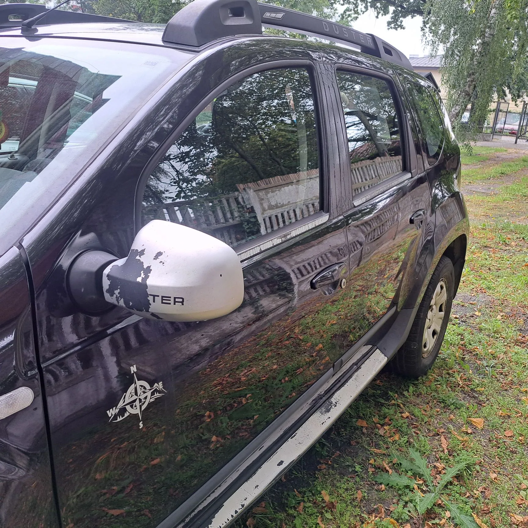 Zbliżenie na prawy bok ciemnofioletowego samochodu marki Dacia Duster. Widoczna jest przednia i tylna drzwi oraz lusterko boczne, którego biała obudowa jest mocno uszkodzona i łuszczy się, odsłaniając ciemniejszy kolor pod spodem oraz fragment napisu 'TER'. Na przednich drzwiach, poniżej lusterka, znajduje się naklejka z motywem kompasu. Samochód zaparkowany jest na trawiastym podłożu z opadłymi liśćmi.
