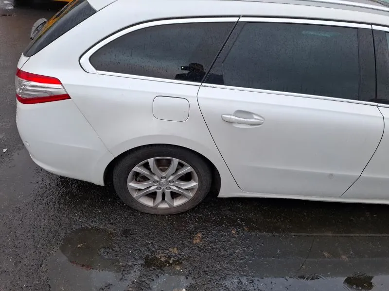 Biały samochód typu kombi widziany z boku i tyłu. Samochód stoi na mokrym asfalcie z kałużami i plamami brudu. Widoczne są czerwone światło tylnego reflektora, przyciemniane szyby, srebrne felgi oraz zamknięty korek zbiornika paliwa. W tle fragment innego pojazdu. Na szybie odbicie osoby trzymającej urządzenie do robienia zdjęć.