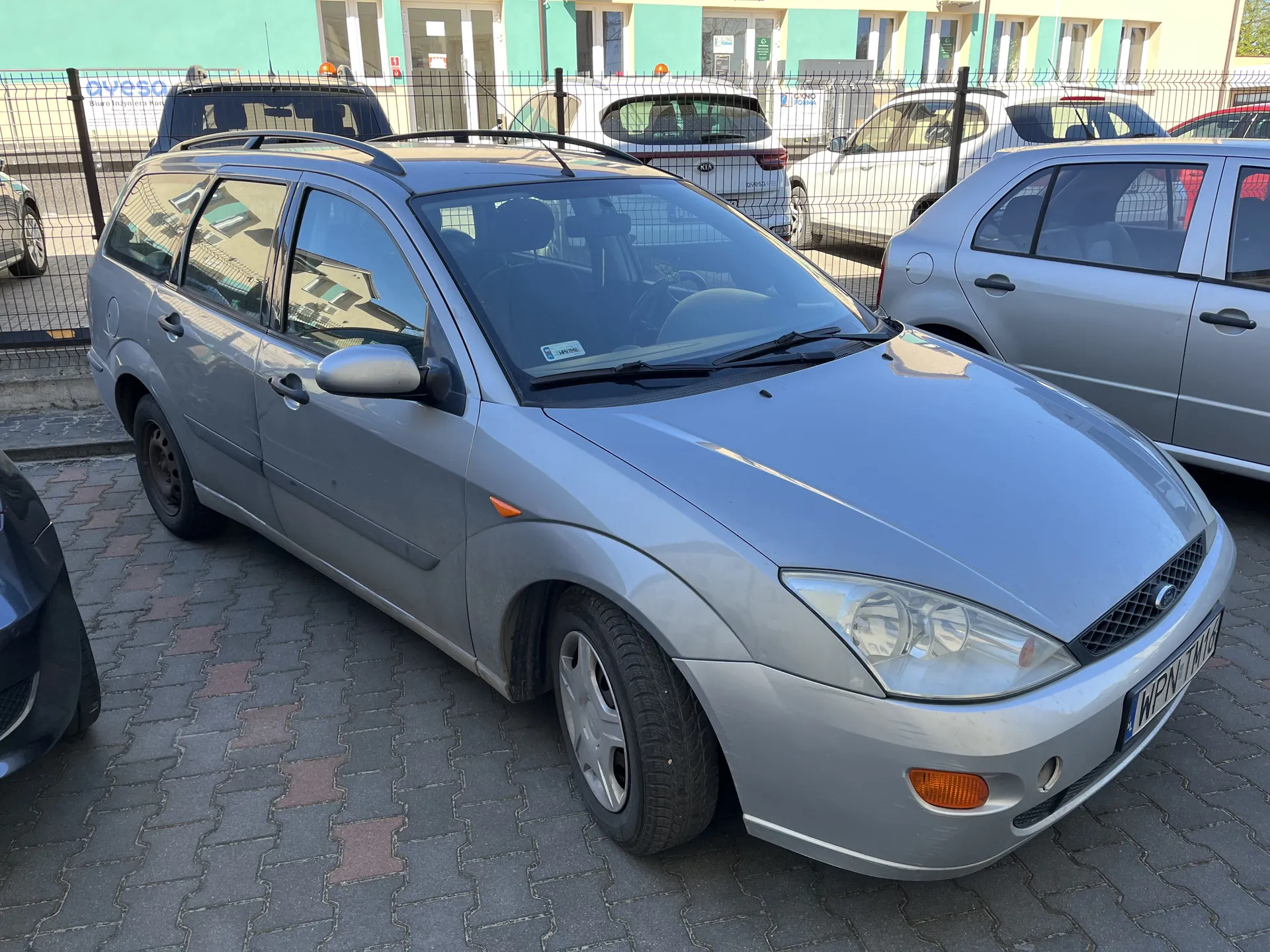 Srebrny samochód typu kombi marki Ford zaparkowany na brukowanej powierzchni. Widoczna tablica rejestracyjna WPN 1M4. W tle inne pojazdy za siatkowym ogrodzeniem oraz budynek z zielono-żółtą elewacją. Na ogrodzeniu widoczny napis aveso. Samochód ma dachowy bagażnik i lusterka boczne.