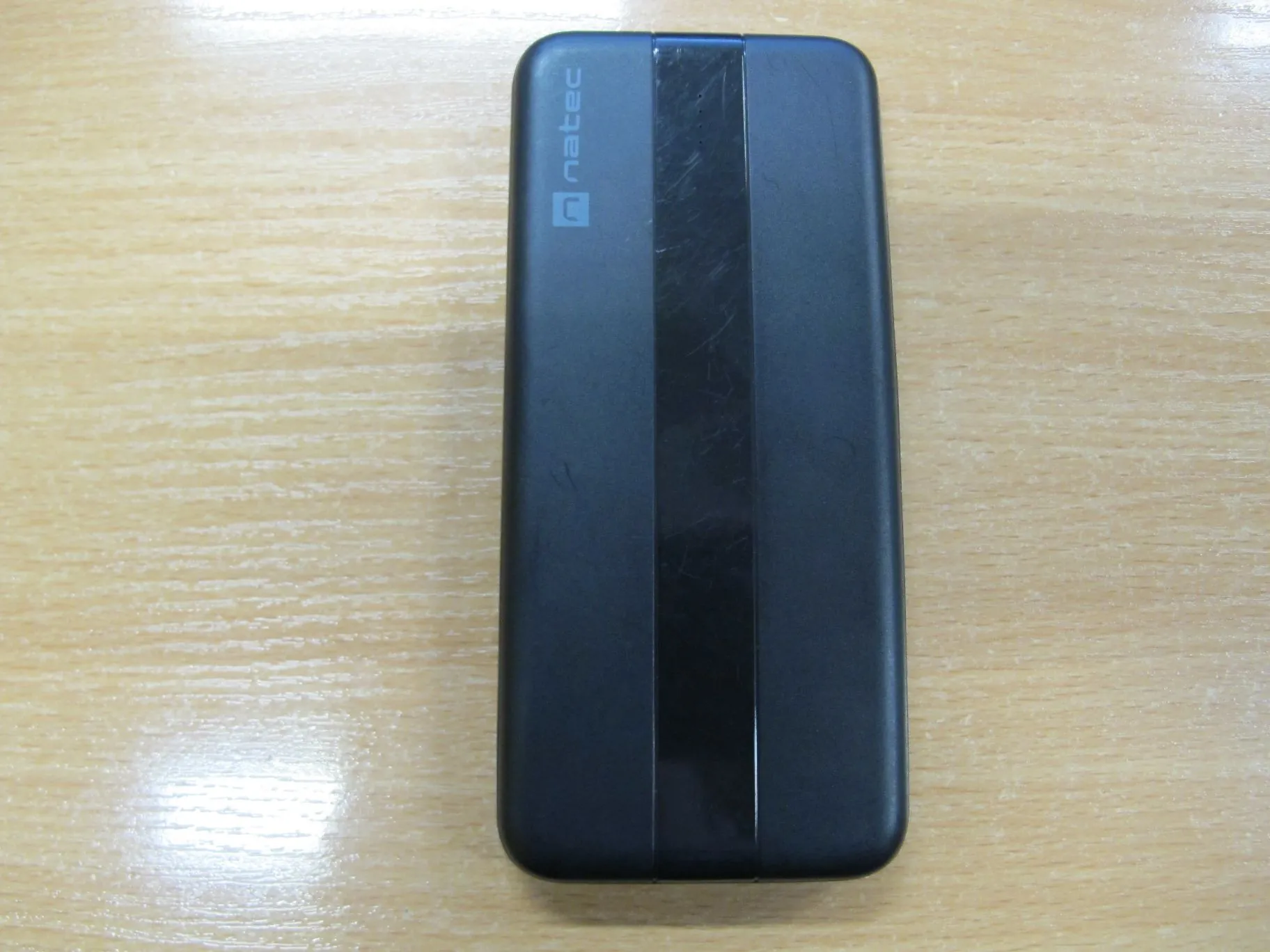Czarny, prostokątny powerbank marki natec leżący na jasnym, drewnianym blacie. Na górnej części urządzenia widoczne jest pionowe logo i napis 'natec'. Na froncie znajduje się szeroki, błyszczący, czarny pas biegnący wzdłuż całego urządzenia.