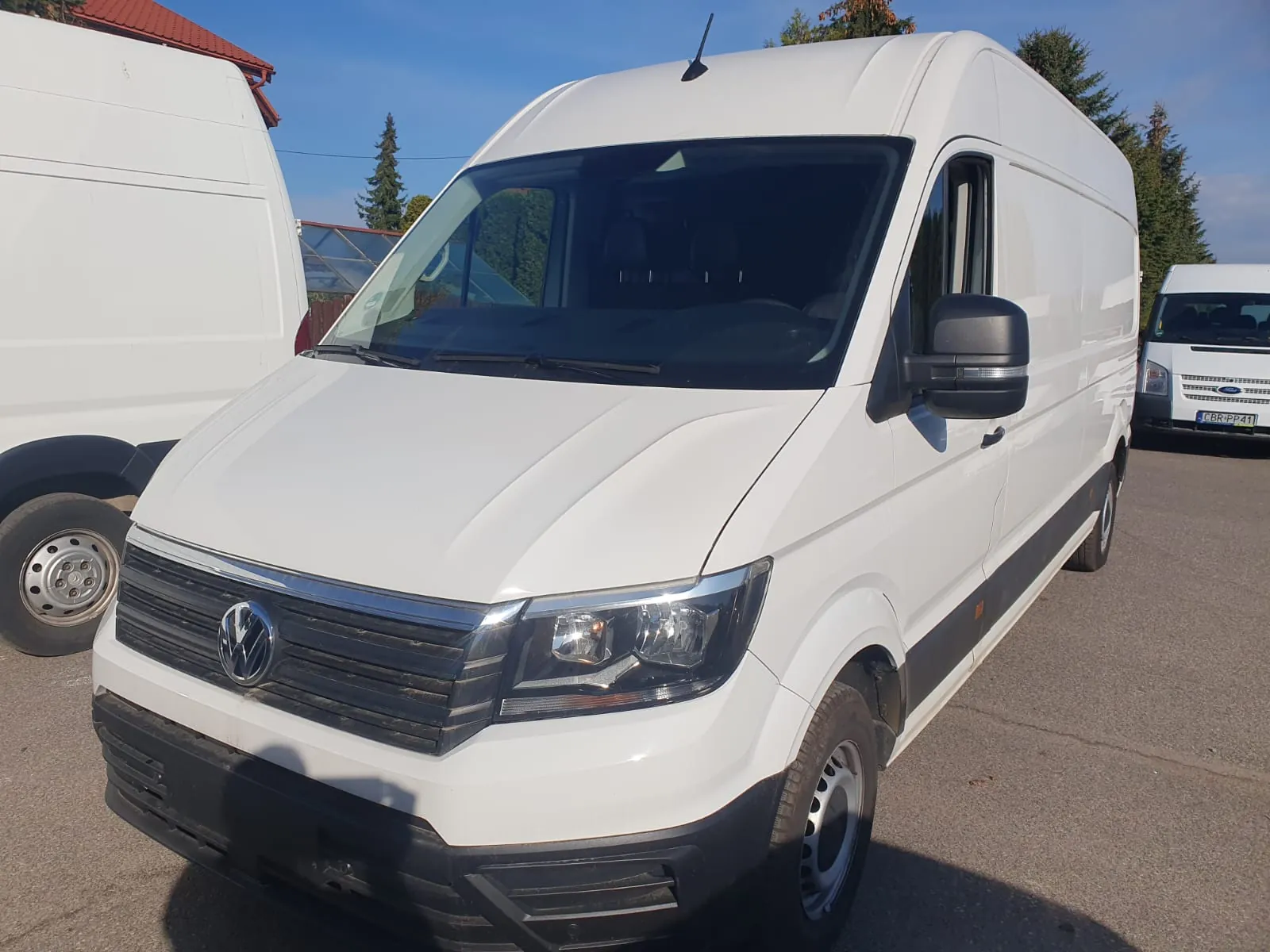 Biały samochód dostawczy marki Volkswagen Crafter zaparkowany na zewnątrz. Pojazd jest widoczny z przodu i z boku. W tle widoczny jest fragment innego białego vana po lewej stronie oraz kolejny biały samochód dostawczy po prawej stronie z widoczną tablicą rejestracyjną CBR PP 41. Niebo jest bezchmurne i niebieskie.