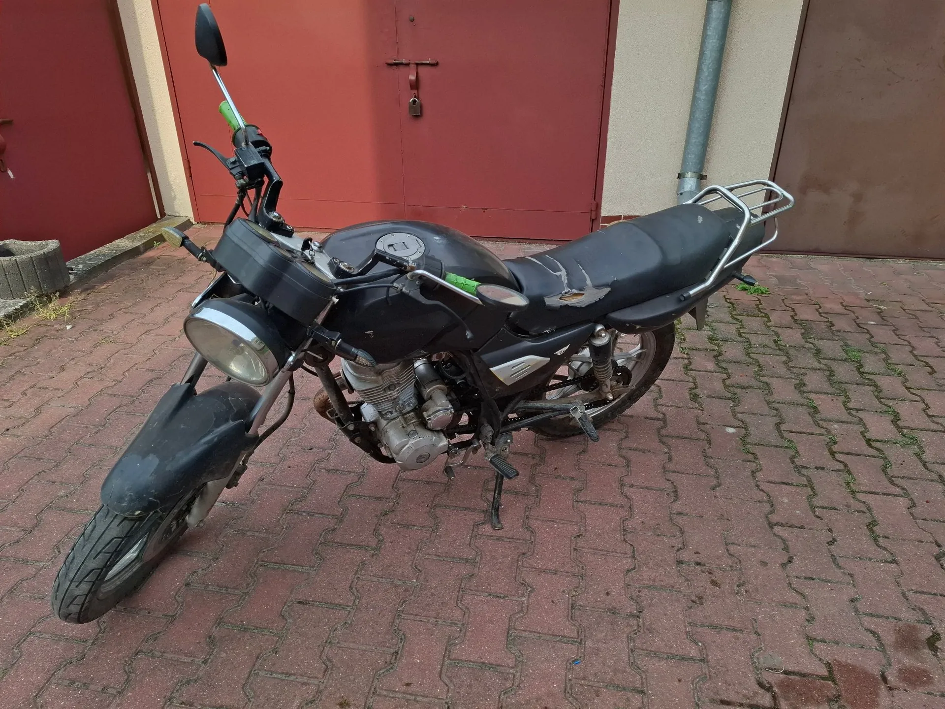 Czarny motocykl zaparkowany na kostce brukowej przed czerwonymi drzwiami garażowymi. Pojazd jest ustawiony pod kątem, widoczny z lewej strony. Ma okrągły reflektor, czarny bak i siedzenie z widocznymi uszkodzeniami materiału. Z tyłu znajduje się metalowy bagażnik. Motocykl stoi na bocznej stopce.
