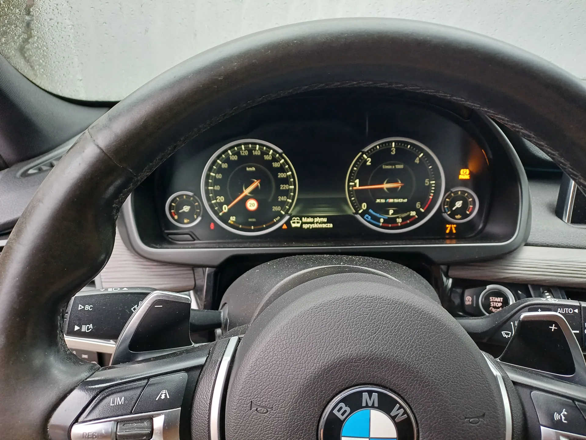 Wnętrze samochodu z widocznym kierownicą marki BMW. Na środku kierownicy znajduje się logo BMW. Przed kierownicą panel przyrządów z dwoma głównymi wskaźnikami: po lewej prędkościomierz (0 km/h) z czerwonym wskaźnikiem, po prawej tachometr (ok. 1 tys. obr./min). W centralnej części wyświetlacza widoczny napis 'Mało płynu spryskiwacza' oraz ikona ostrzegawcza. Obok wskaźników mniej liczne wskaźniki paliwa i temperatury. Na kierownicy przyciski sterujące, w tym 'LIM', 'RES', 'BC' oraz przycisk 'START STOP'. W prawym górnym rogu panelu świeci pomarańczowa ikona 'PARK' i temperatura 7°C.