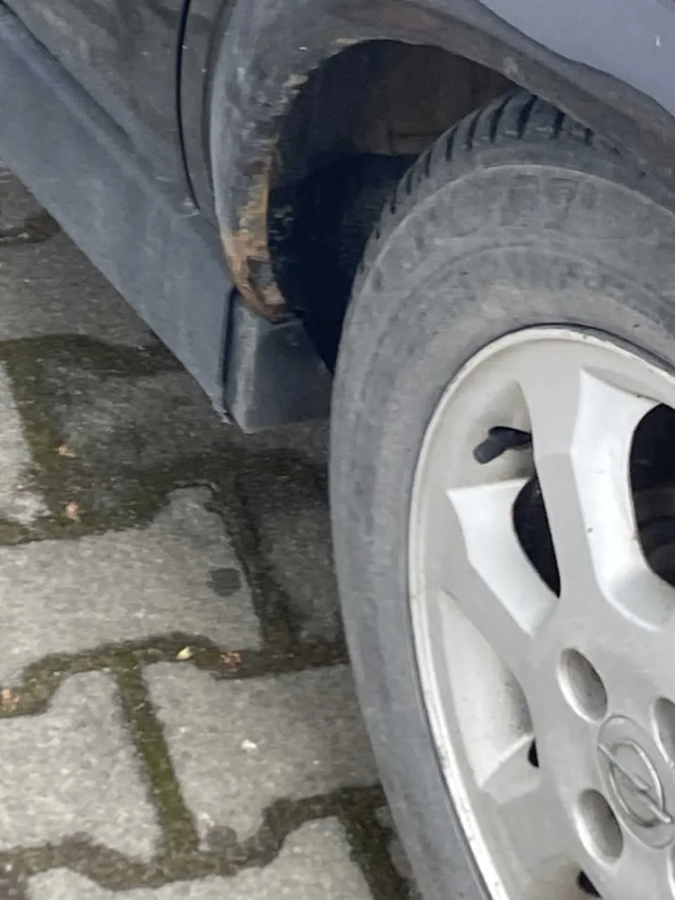 Zbliżenie na przednie koło szarego samochodu z srebrną felgą i oponą. Widoczna jest rdza na zderzaku oraz fragment nawierzchni z betonowych płyt z mchem rosnącym w szczelinach.