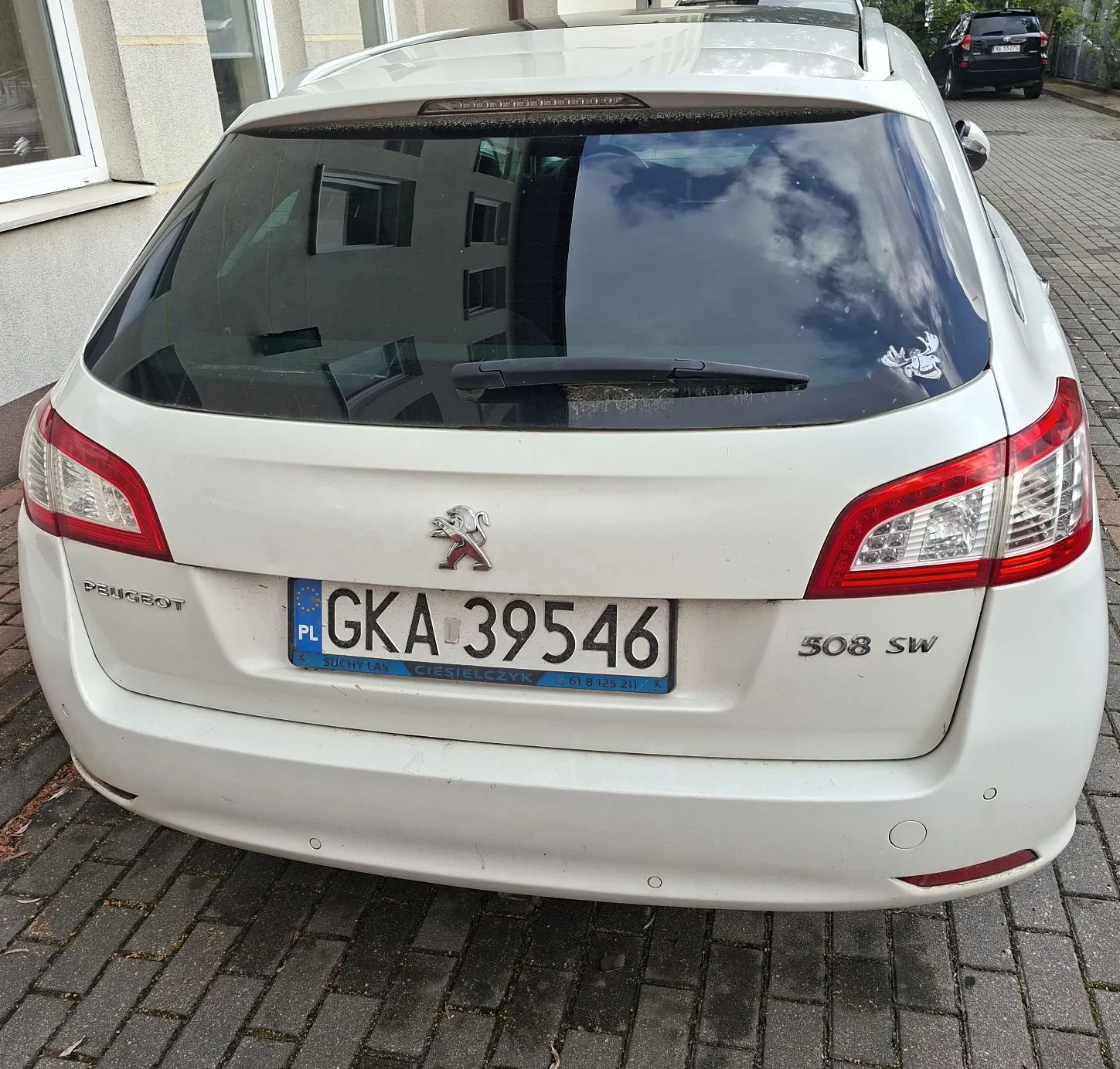Tylny widok białego samochodu marki Peugeot 508 SW. Na klapie bagażnika znajduje się logo marki oraz tablica rejestracyjna o numerze GKA 39546 z oznaczeniem PL. Poniżej tablicy widnieje napis: 'BUCHY LAS CIEŚLIELCZYK 01 8 129 231'. Po prawej stronie znajduje się oznaczenie modelu '508 SW'. Samochód zaparkowany jest na kostce brukowej przed jasnym budynkiem.