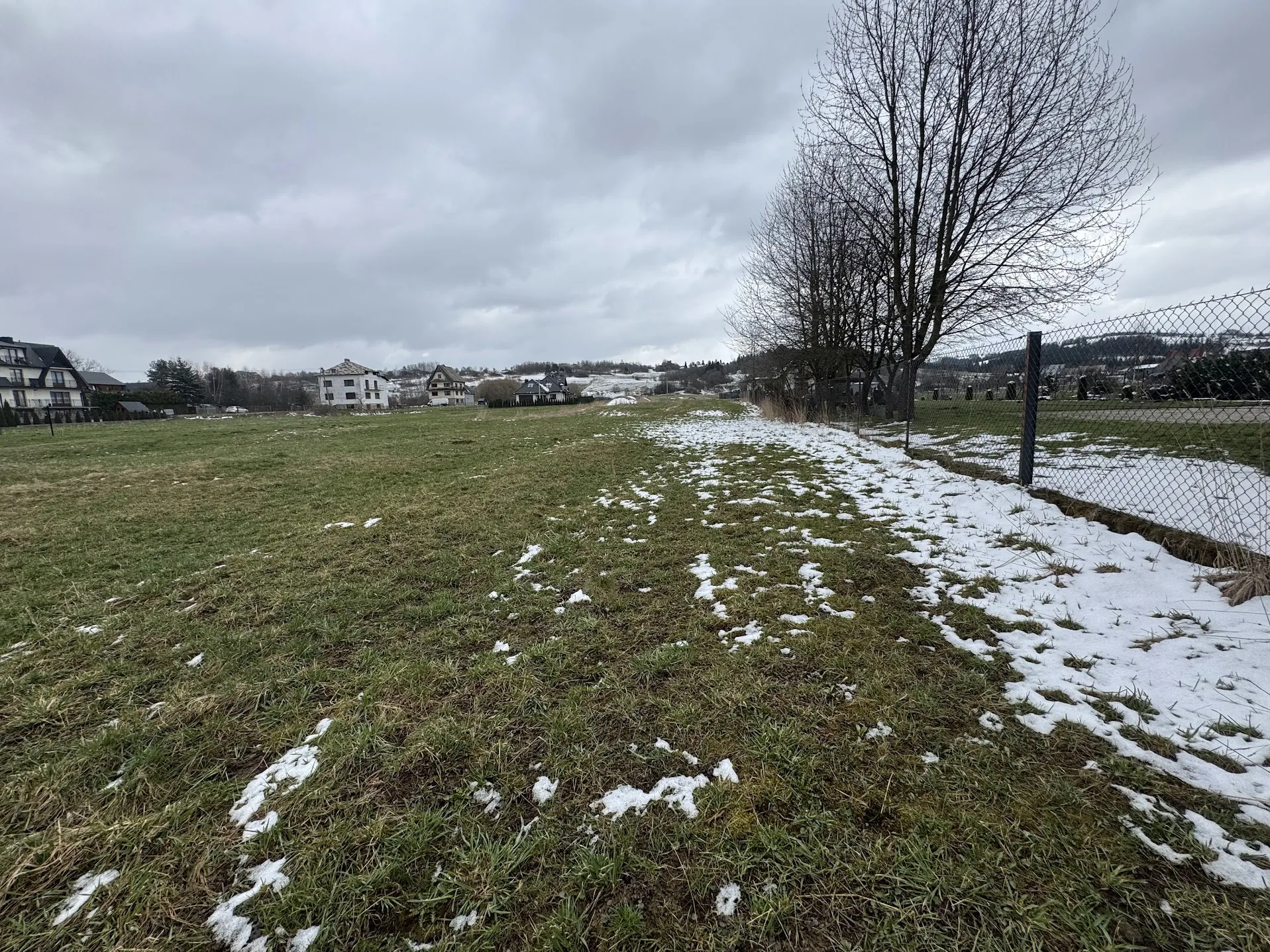 Pole trawiaste z resztkami śniegu, po prawej stronie siatka ogrodzeniowa. W tle domy, bezlistne drzewa i zachmurzone niebo. Horyzont jest poziomy, a obiekty są prawidłowo ustawione.