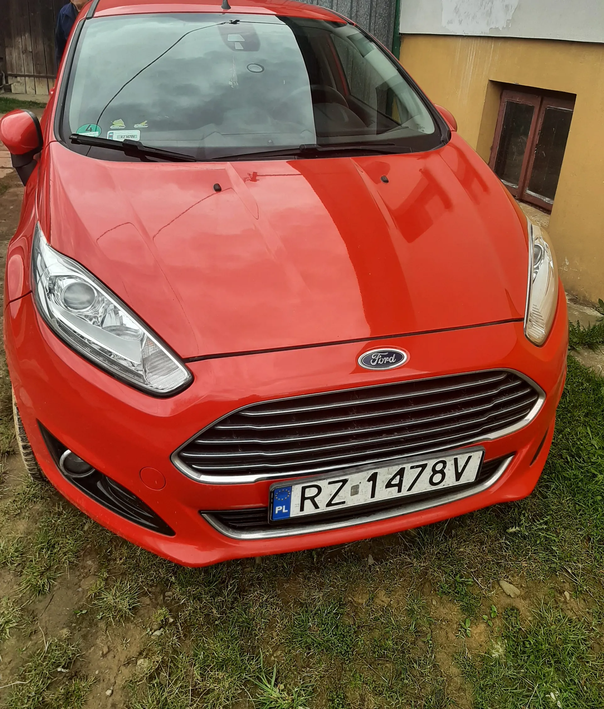 Czerwony samochód marki Ford z przodu, z widocznym logo na atrapie chłodnicy. Tablica rejestracyjna z polskim oznaczeniem PL i numerem RZ 1478V. Na przedniej szybie znajdują się naklejki, w tym zielona z cyfrą 4. Samochód stoi na trawie i ziemi, w tle żółty budynek z brązowym oknem. Niebo jest zachmurzone.