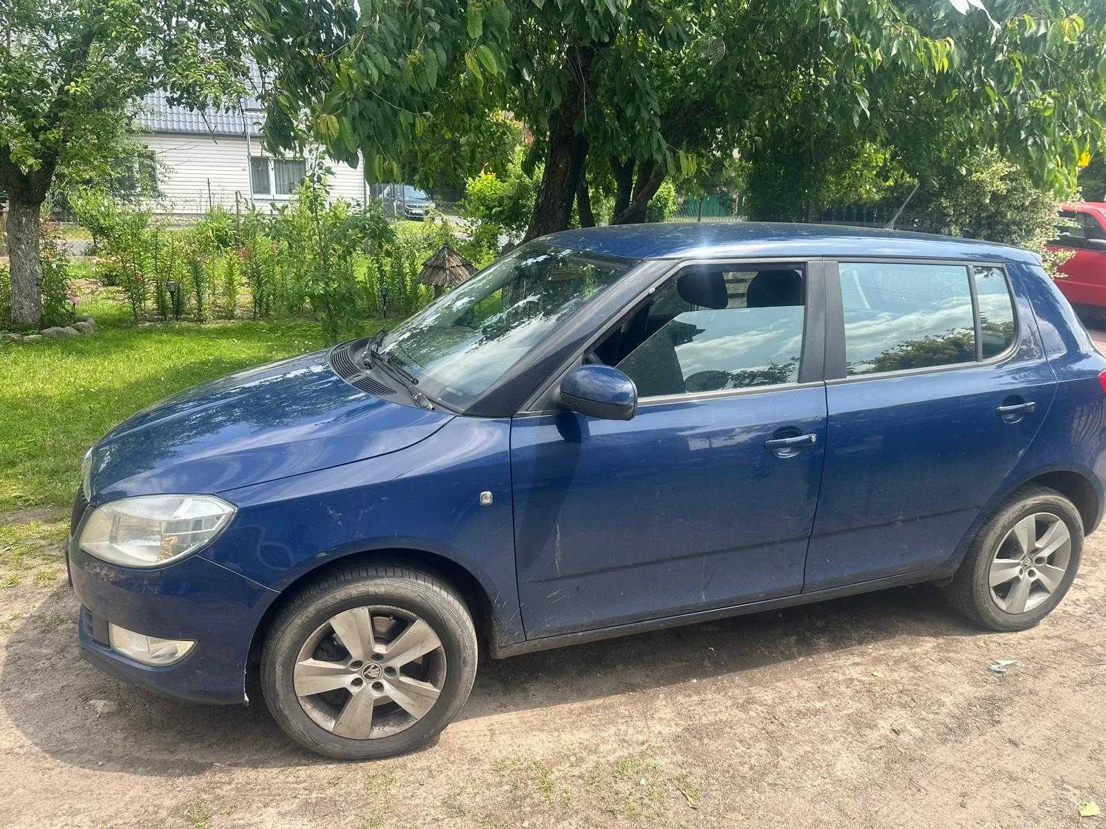Niebieski samochód osobowy typu hatchback zaparkowany na podwórku. Pojazd ma czyste szyby, srebrne felgi i widoczne ślady brudu na karoserii. W tle zielona trawa, drzewa, biały dom z szarą dachówką oraz fragment czerwonego samochodu po prawej stronie. Na podwórku rosną krzewy i widoczny jest mały drewniany daszek.