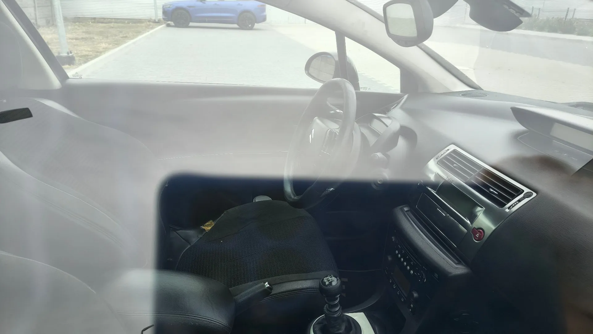 Widok z wnętrza samochodu na deskę rozdzielczą, kierownicę oraz przedni fotel kierowcy. Przez przednią szybę widoczny jest fragment jasnej drogi oraz zaparkowany w oddali niebieski samochód.