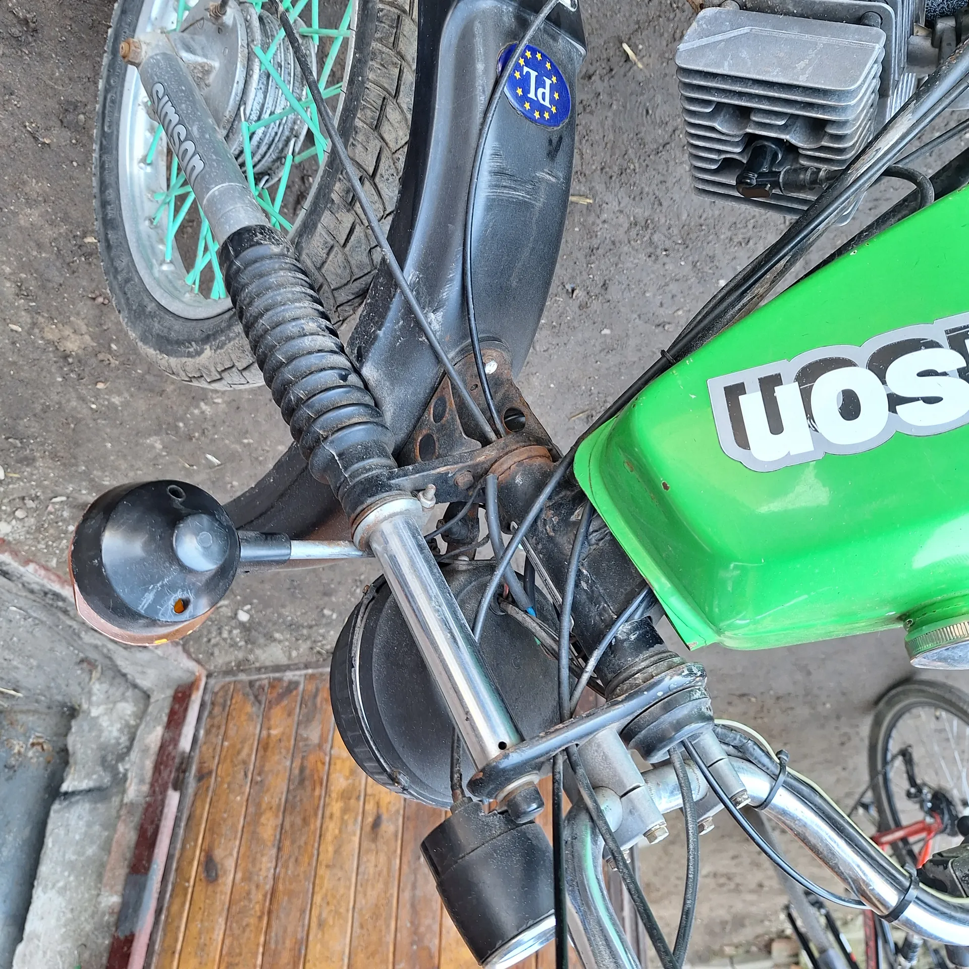 Zbliżenie na fragment motocykla: zielony zbiornik paliwa z częściowo widocznym napisem "WOS", czarna kierownica z gumową obudową, przednie koło z zielonymi promieniami i oponą, reflektor, przewody elektryczne oraz niebieska naklejka z napisem "PL" i żółtymi gwiazdkami. W tle widoczne drewniane drzwi i betonowa podłoga. Fragment innego roweru w lewym dolnym rogu.