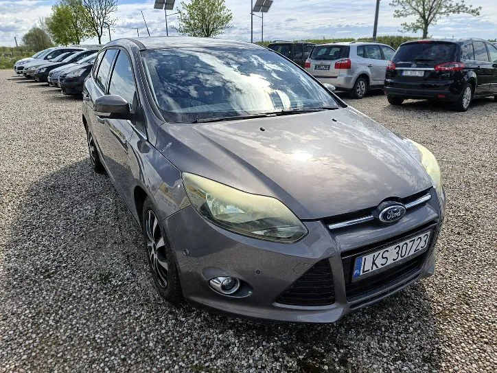 Szary samochód marki Ford zaparkowany na żwirowym placu. Pojazd jest ustawiony pod kątem, widoczny z przodu i z lewej strony. Na przednim zderzaku znajduje się tablica rejestracyjna o numerze LKS 30723. W tle widoczne są inne zaparkowane samochody oraz drzewa pod jasnym, lekko zachmurzonym niebem.