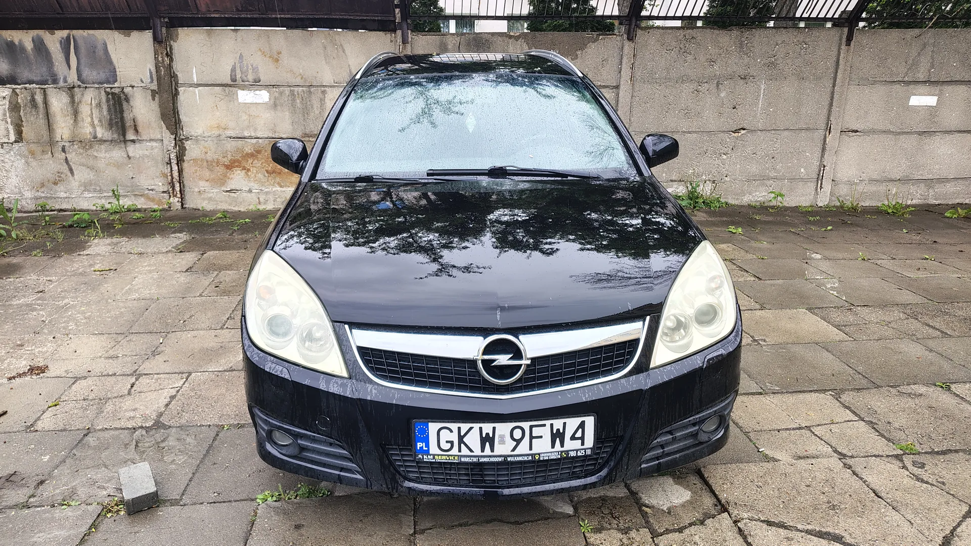 Czarny samochód marki Opel z przodu, z widocznym logo na atrapie chłodnicy. Tablica rejestracyjna PL GKW 9FW4 z dodatkowym napisem poniżej. Samochód stoi na brukowanej powierzchni przed betonową ścianą z metalowym ogrodzeniem na górze. Na ścianie widoczne są plamy i ślady zużycia. W tle rosną pojedyncze rośliny między płytami.