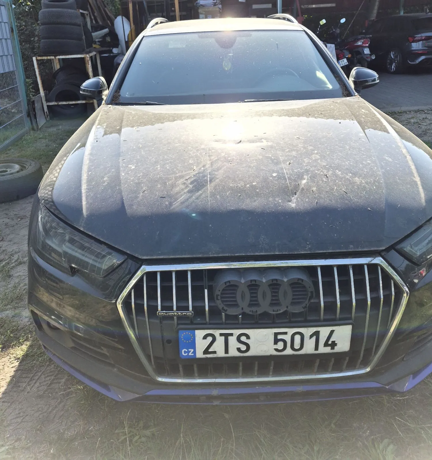 Widok z przodu na ciemny, zakurzony samochód marki Audi z zamontowanym na grillu czeskim numerem rejestracyjnym 2TS 5014. Pojazd znajduje się na zewnątrz, w tle widoczne są opony i inne samochody.
