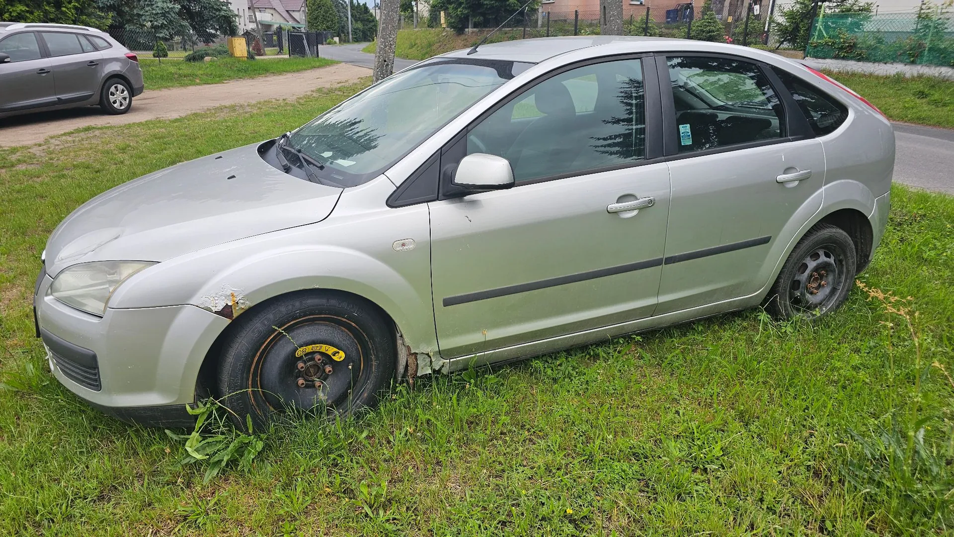 Srebrny samochód osobowy marki Ford Focus zaparkowany na trawie. Pojazd jest uszkodzony: przednie lewe koło jest całkowicie zapadnięte i wbite w ziemię, a na przednim błotniku widoczne są wgniecenia i odpryski lakieru. W tle widać fragment innego, szarego samochodu oraz zielone otoczenie z drzewami i niskim ogrodzeniem.