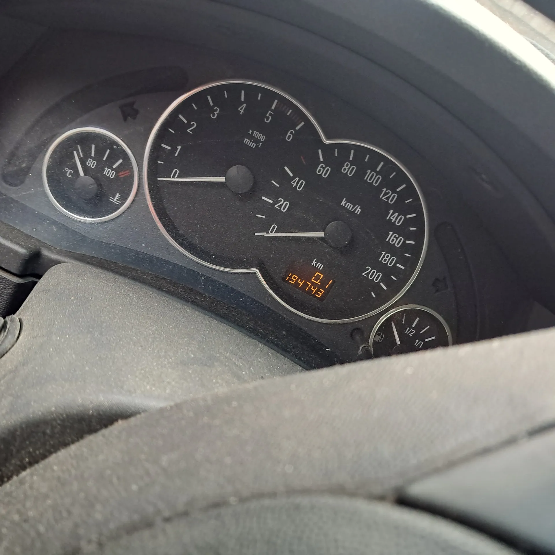 Zbliżenie na desce rozdzielczej samochodu z licznikami. W centrum znajduje się prędkościomierz do 200 km/h, obok tachometr do 7 (x1000 obr./min). Wyświetlacz cyfrowy pokazuje przebieg 19473 km. Po lewej stronie wskaźnik paliwa, po prawej termometr. Fragment kierownicy widoczny u góry.