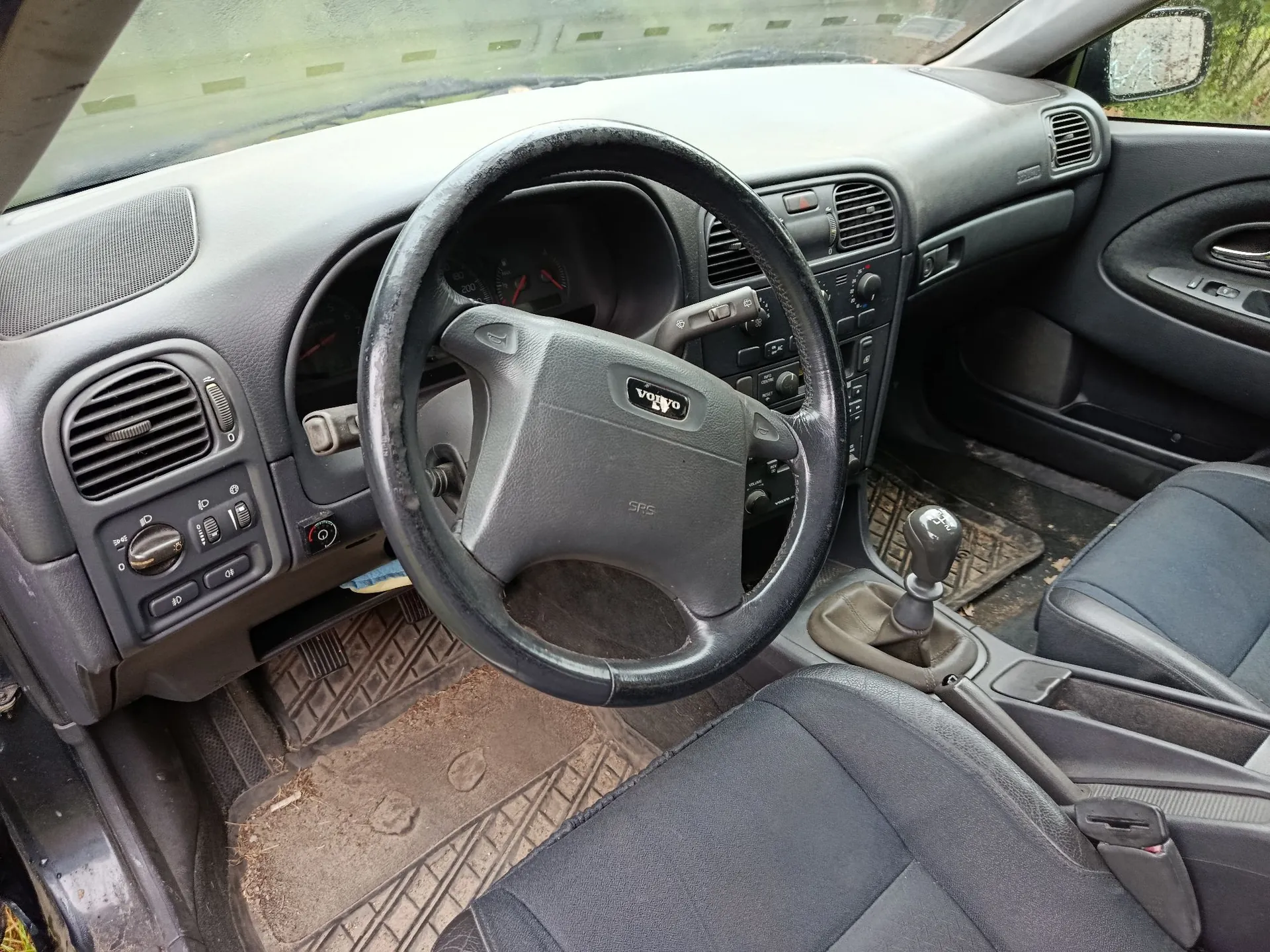 Wnętrze samochodu marki Volvo z perspektywy kierowcy. Widoczna jest czarna kierownica z logo VOLVO i oznaczeniem SRS, deska rozdzielcza z kratkami nawiewów, przełącznikami świateł oraz panel sterowania radiem i klimatyzacją. W tle widać ciemną tapicerkę foteli oraz gumowe dywaniki na podłodze.