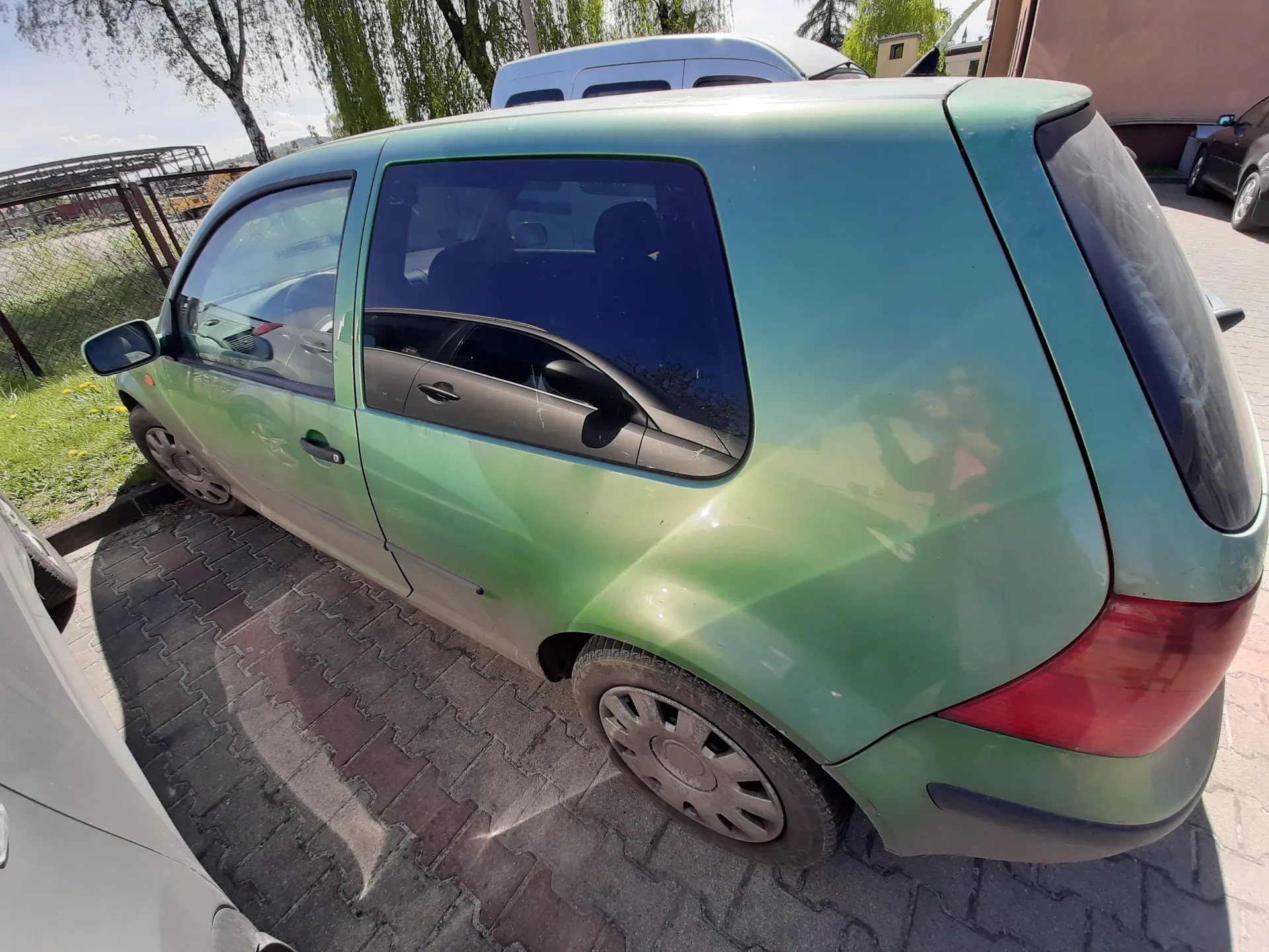 Zanieczyszczony zielony samochód typu hatchback zaparkowany na brukowanej powierzchni. W tle widoczne są inne pojazdy, drzewa, ogrodzenie i budynki. Na karoserii odbijają się sylwetki osób i otoczenia.