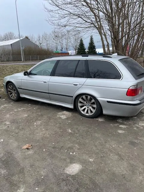 Srebrny samochód typu kombi zaparkowany na betonowej powierzchni. W tle widoczne są bezlistne drzewa, niewielkie budynki, iglaste drzewa oraz pochmurne niebo. Samochód ma czarne felgi i ciemne szyby. Na ziemi leżą rozrzucone liście i plamy.