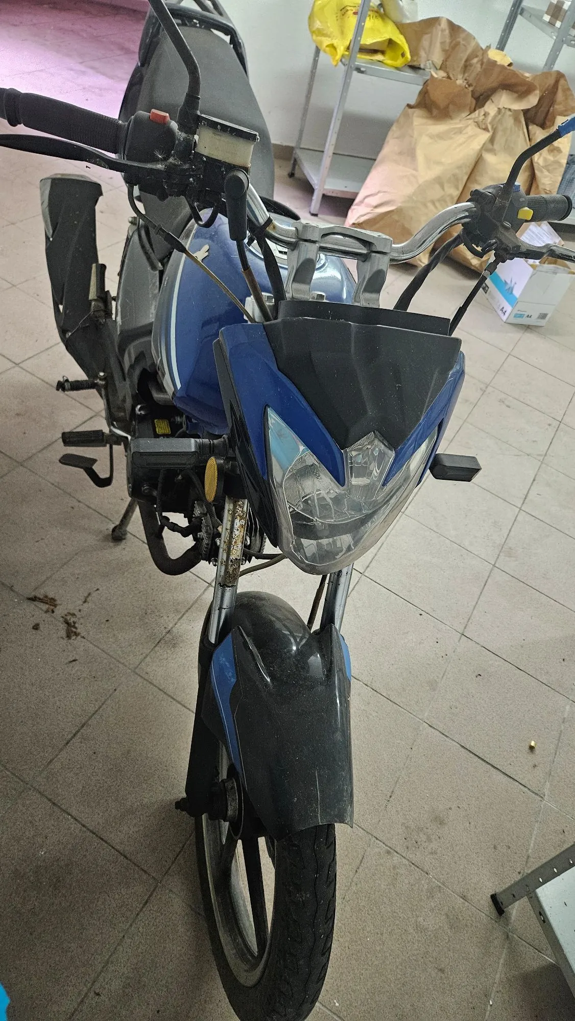 Niebieski motocykl zaparkowany w pomieszczeniu z szarą płytkową podłogą. Widoczny jest przód pojazdu z reflektorem, kierownicą oraz przednim kołem z czarnym błotnikiem. W tle znajdują się metalowe regały z brązowymi papierowymi torbami i białym pudełkiem.