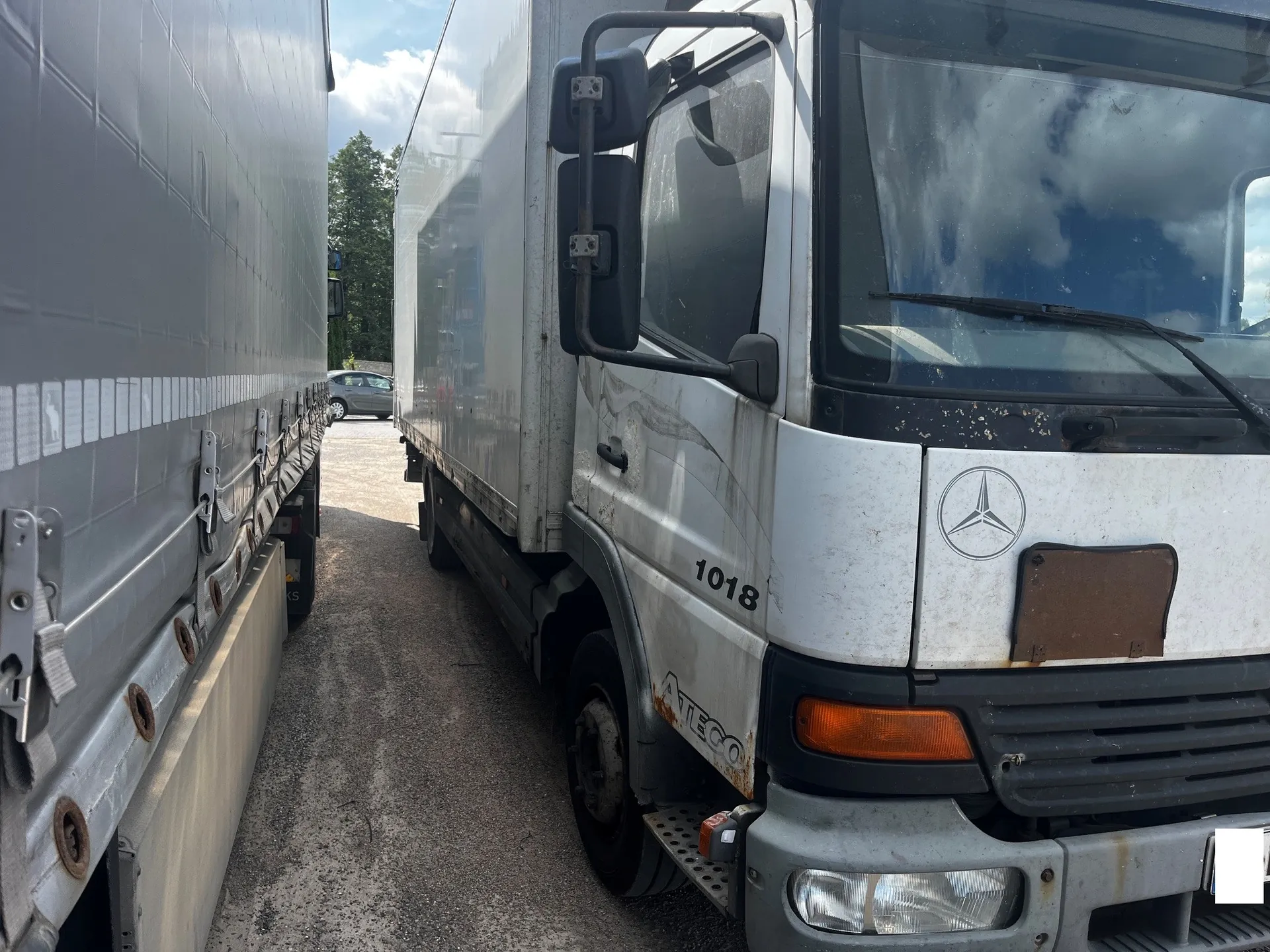 Zbliżenie na przód i bok białej ciężarówki marki Mercedes-Benz Atego, model 1018. Pojazd jest częściowo zabrudzony, widoczne jest logo marki na masce oraz napis '1018' i 'ATEGO' na drzwiach. Po lewej stronie kadru znajduje się fragment białej naczepy innego pojazdu. W tle widoczny jest parking, srebrny samochód osobowy oraz drzewa pod zachmurzonym niebem.