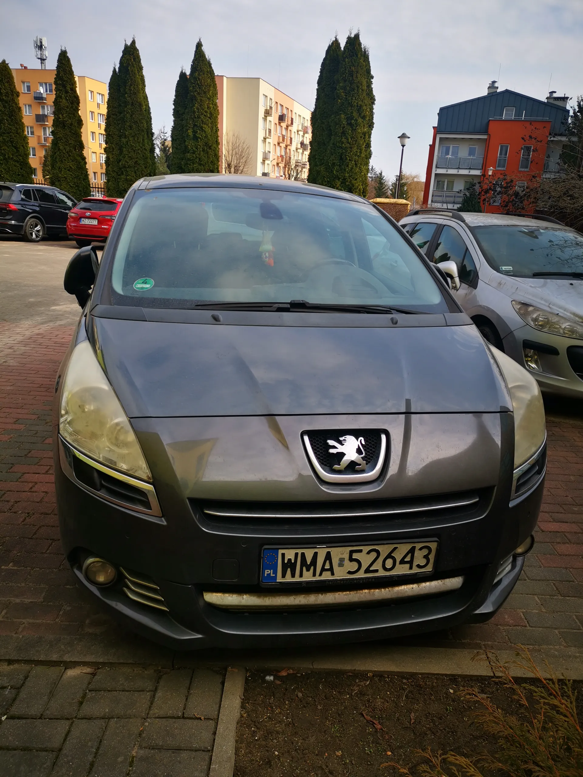 Przód szarego samochodu marki Peugeot z tablicą rejestracyjną WMA 52643 (PL) na tle parkingu z innymi pojazdami, budynkami mieszkalnymi i wysokimi drzewami. Na przedniej szybie widoczna zielona naklejka z cyfrą 4.