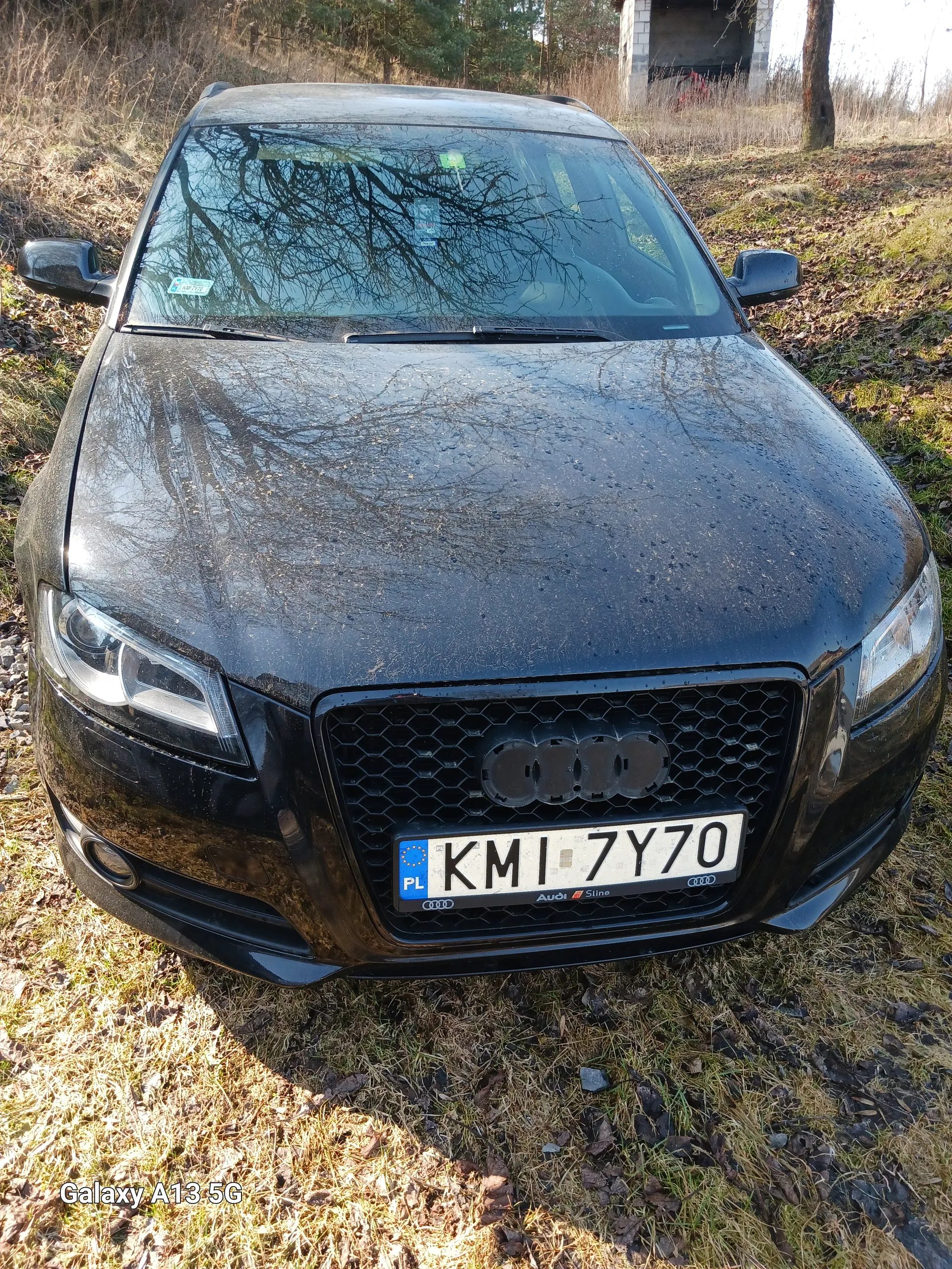 Czarny samochód marki Audi z przodu, tablica rejestracyjna KMI 7Y70 z oznaczeniem PL i niebieskim pasem z gwiazdkami. Samochód zaparkowany na trawie z suchymi liśćmi. W tle widoczny budynek i drzewa. Na szybie przedniej odbicia drzew. Po prawej stronie obrazu widoczny pionowy napis 'Galaxy A13 5G'.