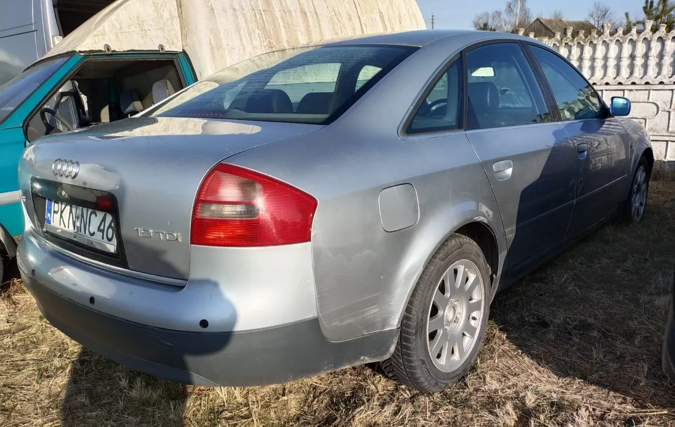 Srebrny samochód Audi sedan z widocznym tyłem, tablicą rejestracyjną PK NNC 46 oraz napisem 1.9 TDI. Po lewej stronie częściowo widoczny turkusowy furgon z otwartymi drzwiami. Tło: sucha trawa, betonowa konstrukcja i kamienny płot. Samochód stoi na poziomym terenie w świetle dziennym.