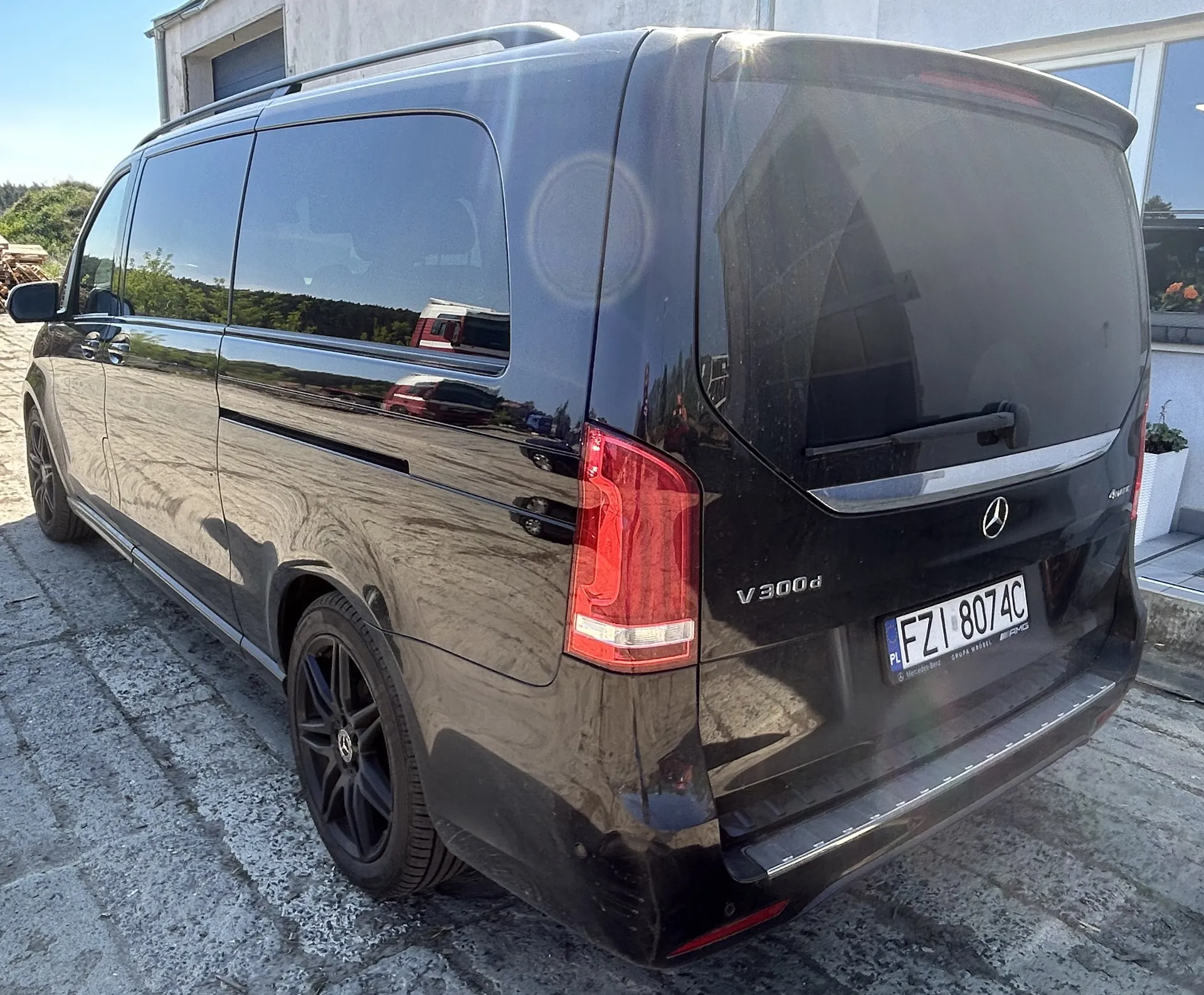 Czarny samochód dostawczy Mercedes-Benz V300d zaparkowany na brukowanej powierzchni. Widoczny tył pojazdu z tablicą rejestracyjną PL FZI 8074C, logo Mercedes-Benz oraz napisem V300d. W tle budynek z garażem i zielonymi krzewami, jasne niebo.