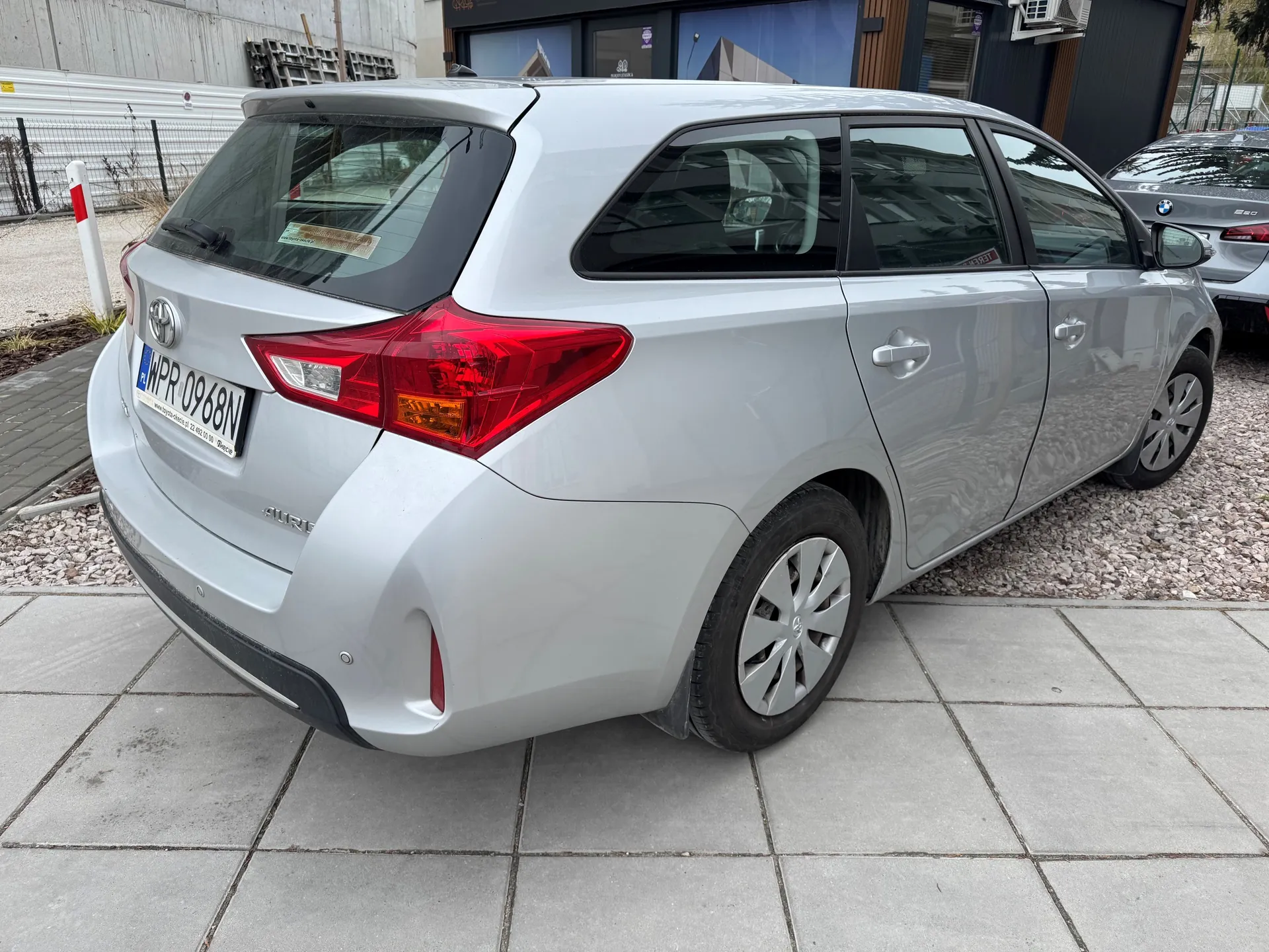 Srebrny samochód osobowy typu kombi marki Toyota Auris zaparkowany na chodniku z kostki. Widoczny tył pojazdu z czerwonymi światłami, tablicą rejestracyjną WPR 0968N oraz logo marki. W tle drugi samochód (BMW), budynek z dużymi szybami, ogrodzenie i materiały budowlane. Podłoże składa się z kostki chodnikowej i żwiru.