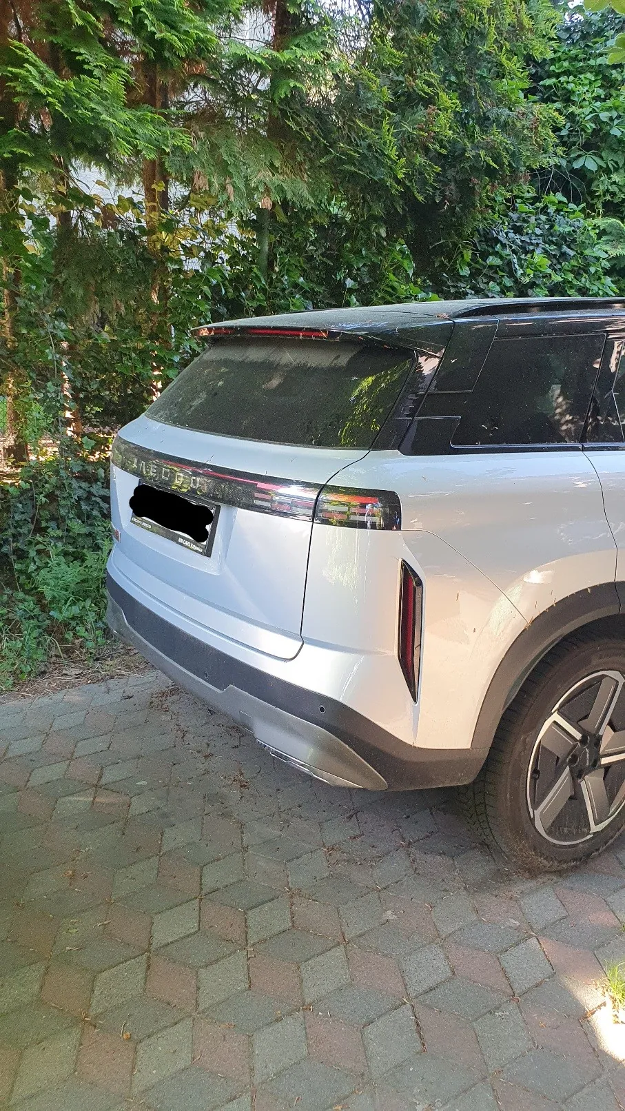 Tylna część białego samochodu marki Range Rover zaparkowanego na kostce brukowej. Pojazd znajduje się na tle gęstych, zielonych drzew i krzewów. Tablica rejestracyjna jest zakryta czarnym polem.