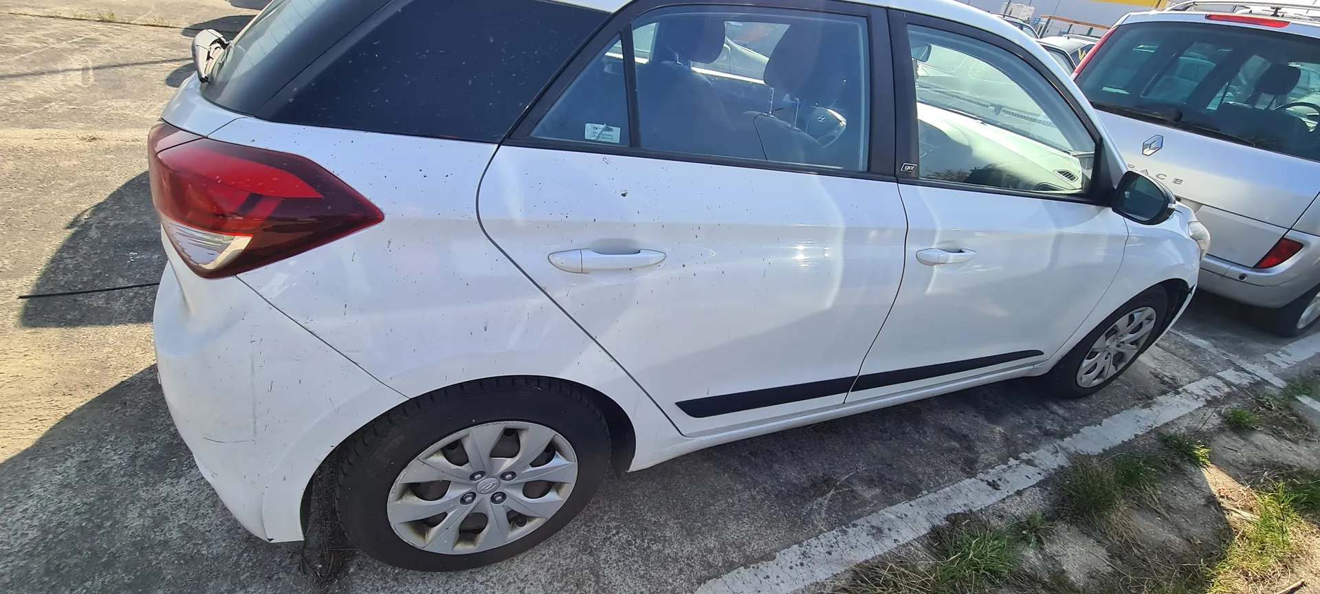 Biały samochód osobowy typu hatchback zaparkowany na betonowym placu parkingowym. Widoczne są czerwone światła tylne, srebrne felgi, czarne listwy boczne oraz zabrudzenia na karoserii. W tle drugi srebrny samochód i fragmenty asfaltu z zielonymi plamami trawy. Słoneczne oświetlenie rzuca cienie na powierzchnię parkingu.