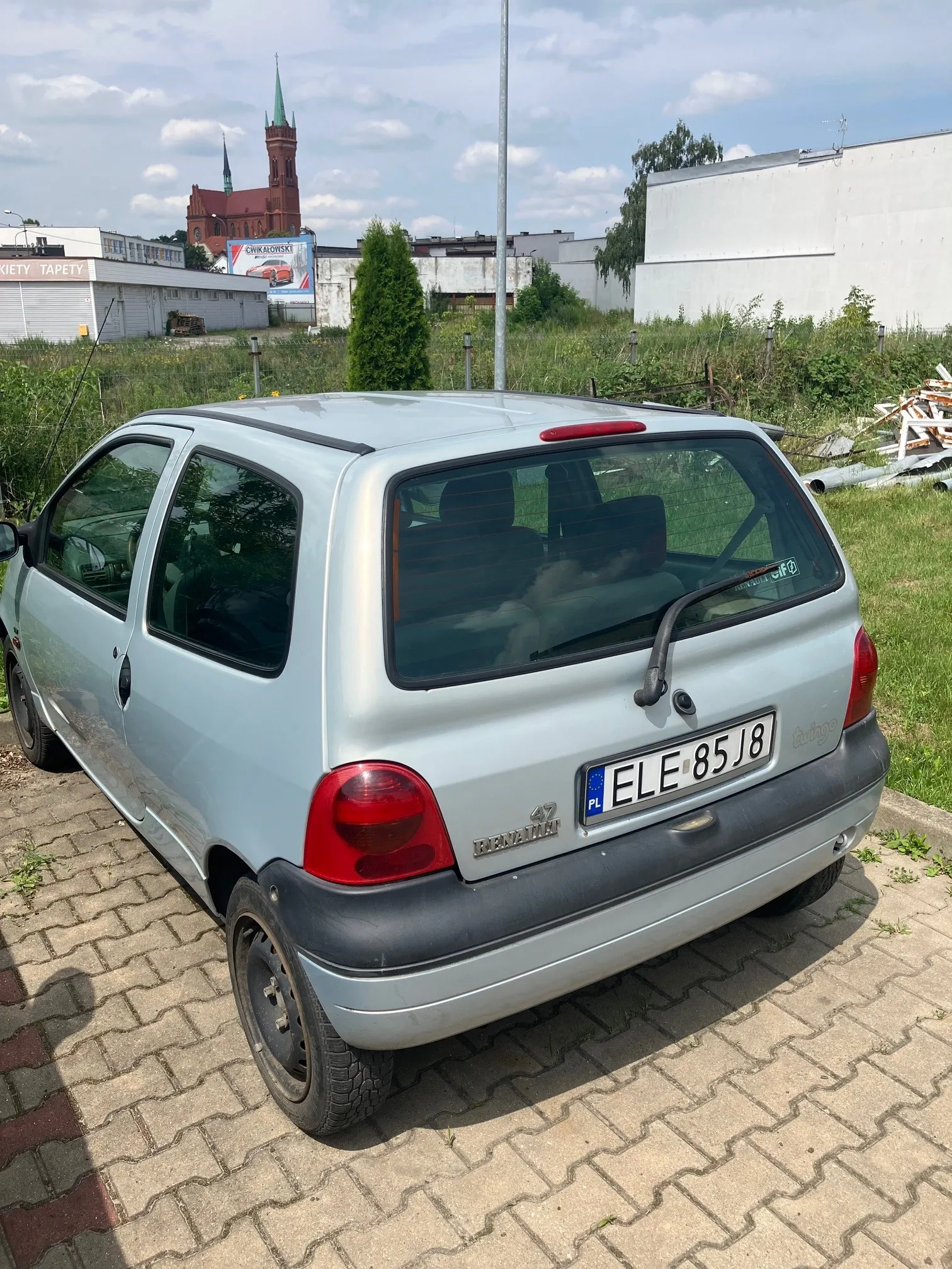 Srebrny samochód osobowy marki Opel Corsa w wersji nadwozia hatchback, zaparkowany tyłem na kostce brukowej. Pojazd posiada polską tablicę rejestracyjną o numerze ELE 85J8. W tle widoczny jest zielony teren, białe budynki przemysłowe oraz czerwona wieża kościoła z zielonym szczytem pod zachmurzonym niebem.