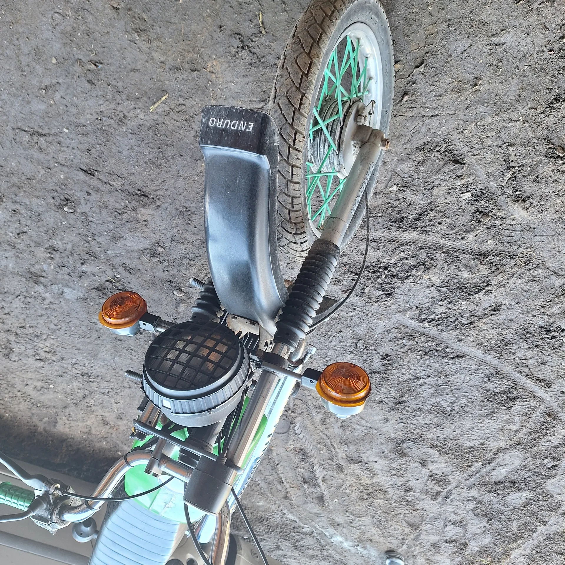 Przednia część motocykla z kołem o zielonych promieniach, czarnym błotnikiem z napisem ENDURO, dwoma pomarańczowymi kierunkowskazami, reflektorem i zielonym zbiornikiem paliwa. Tło stanowi brudna, ziemista powierzchnia z odciskami opon.