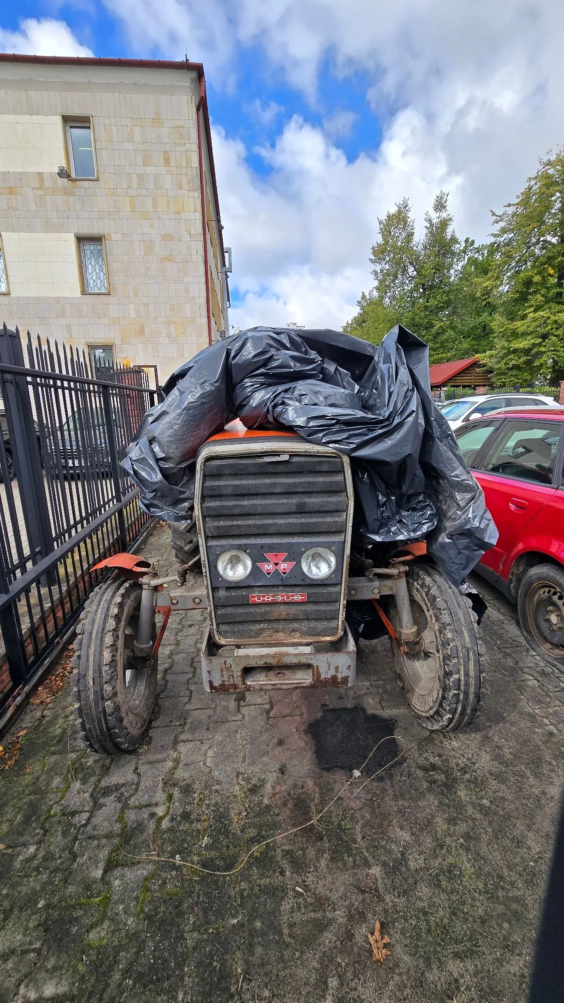 Stary, pomarańczowy traktor marki Ursus, częściowo przykryty czarną foliową plandeką. Pojazd stoi na betonowym podjeździe obok czarnego metalowego ogrodzenia i czerwonego samochodu. Na przodzie traktora widoczny jest napis 'URSUS' oraz logo marki.