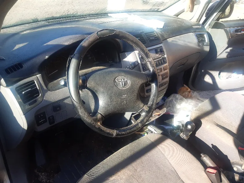 Wnętrze samochodu marki Toyota z widocznym kierownicą z logo, zużytą obiciem kierownicy, deska rozdzielcza z przyciskami i wentylacją, dźwignią zmiany biegów, siedzeniami tapicerowanymi tkaniną, otwartą drzwiami po stronie kierowcy oraz plastikowymi workami na siedzeniu pasażera. Wnętrze wydaje się zabrudzone, a na desce rozdzielczej i siedzeniach widoczne są ślady zużycia. Światło słoneczne wpada przez przednią szybę, rzucając cienie na wnętrze.